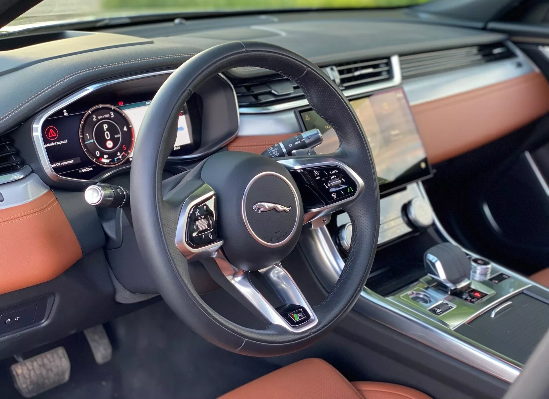 The image displays a close-up view of the 2021 Jaguar XF interior focused on the driver's side from the front passenger seat. The foreground features a modern three-spoke steering wheel with multifunction buttons and the Jaguar emblem. Behind the wheel, a partially visible instrument cluster displays digital information. To the right is the car’s infotainment system on the central dashboard, likely controlling audio, navigation, and various settings. The luxurious interior showcases a soft-touch dashboard with top-stitched leather accents in deep tan, darker tones, metallic accents, and a gear shift lever suggesting automatic transmission and controls for climate and driving modes. High-quality lighting enhances the details.