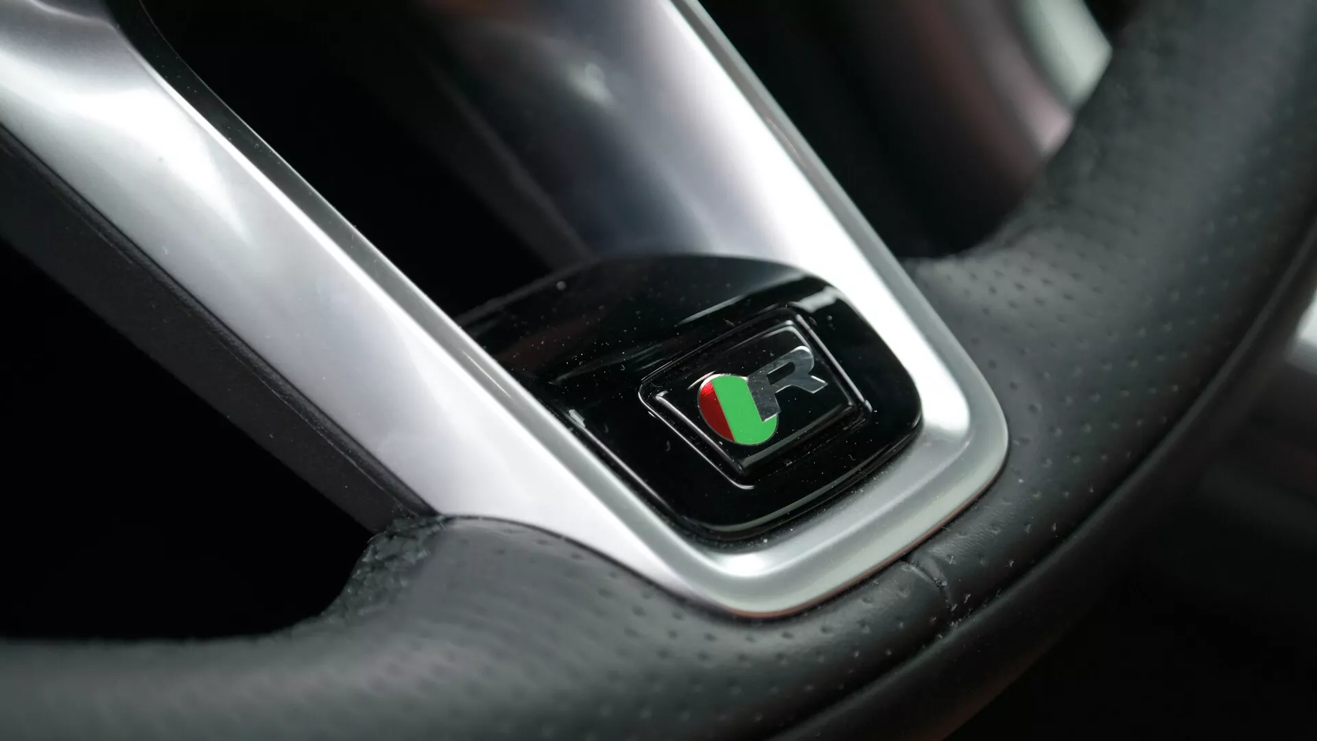Close-up driver's perspective view of the 2021 Jaguar XF steering wheel, featuring a digital display at the center showing a racing flag, suggesting performance mode engaged. The image highlights the luxury leather material with visible stitching and carbon fiber texture, complemented by a chrome accent around the screen, reflecting bright lighting.