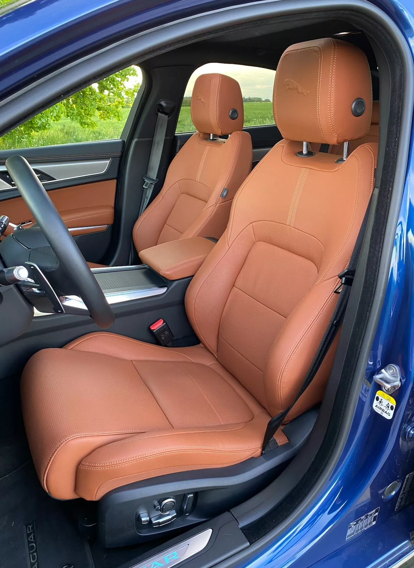 Side-angle view of the luxurious interior of a 2021 Jaguar XF, focusing on tan leather-upholstered driver's and passenger seats with electronic adjustments; partial center console, and side airbag label visible; vibrant blue exterior paint contrasts with the cabin's two-tone color scheme.