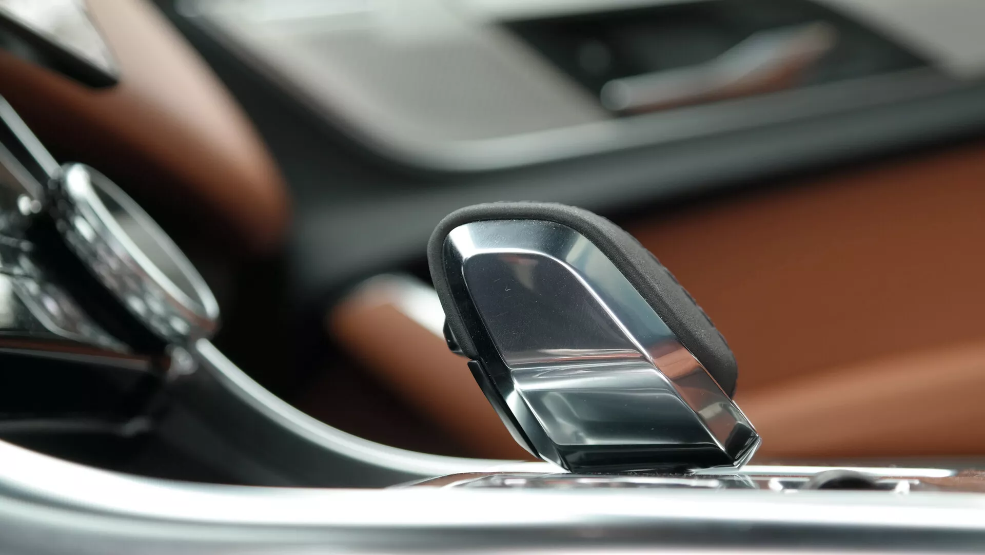 Close-up view of the Jaguar XF 2021 interior, focusing on a sleek joystick or dial-type gear selector. The perspective is slightly above and angled downward, highlighting the gear selector in the foreground. To the left, part of a round control knob related to the infotainment or audio system is visible. The dashboard, finished with luxurious wood trim, is shown behind the gear selector, with a touch screen integrated into the metallic-accented central console in the background. The blurred background emphasizes the gear selector, suggesting a shallow depth of field. This implies the car's automatic transmission and modern infotainment system, indicating a higher-end, luxury vehicle.