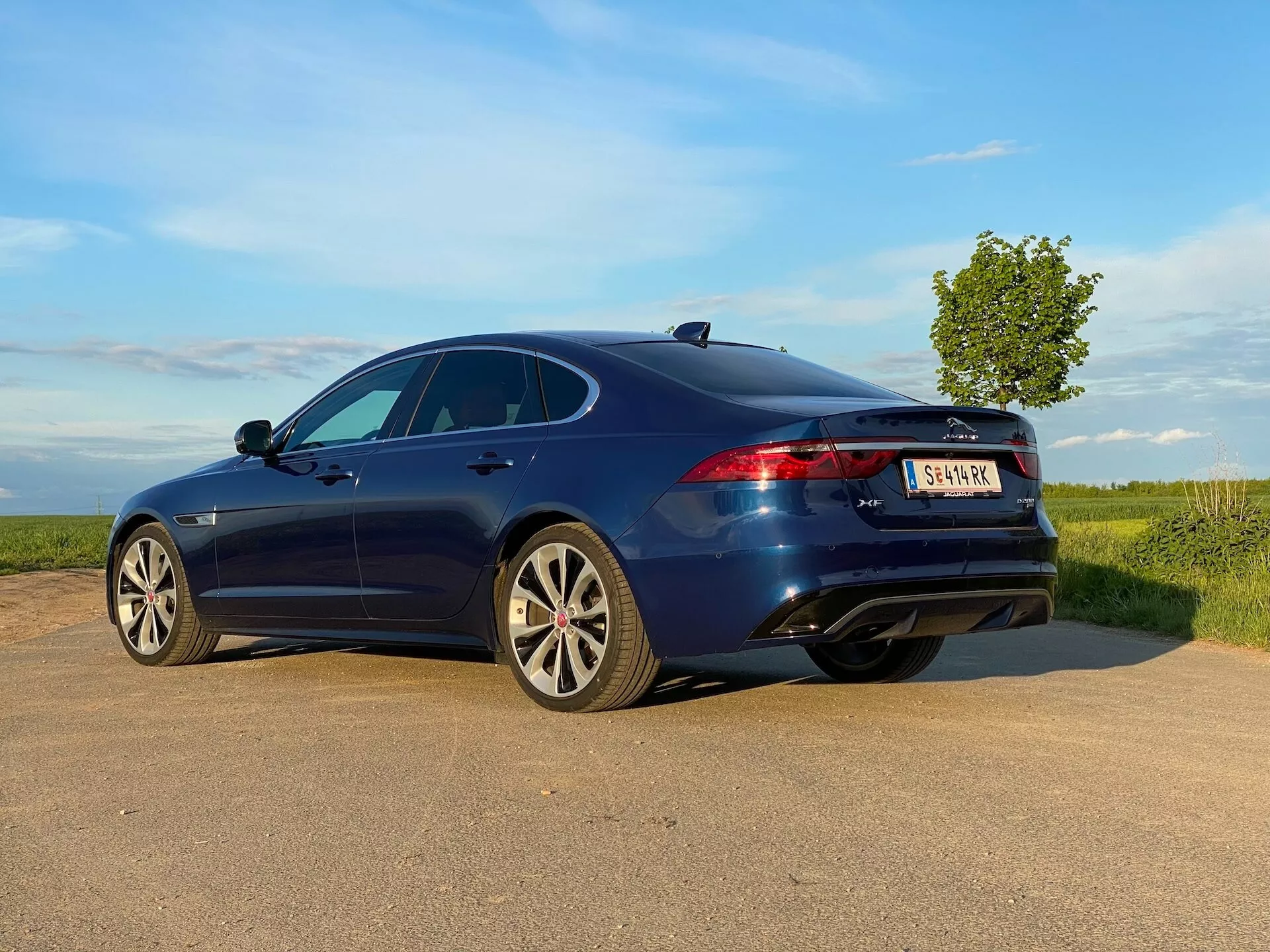 Modern four-door sedan in deep blue seen from a rear three-quarter view on a country road. The vehicle features a sleek, aerodynamic profile with a fastback design and alloy wheels with a multiple-spoke design. The image also shows a sunroof, modern LED taillights, a third brake light, and dual exhaust pipes. The natural, partly cloudy sky in the background suggests an open, rural setting, adding to the dynamic feel of the photograph taken from a low angle.