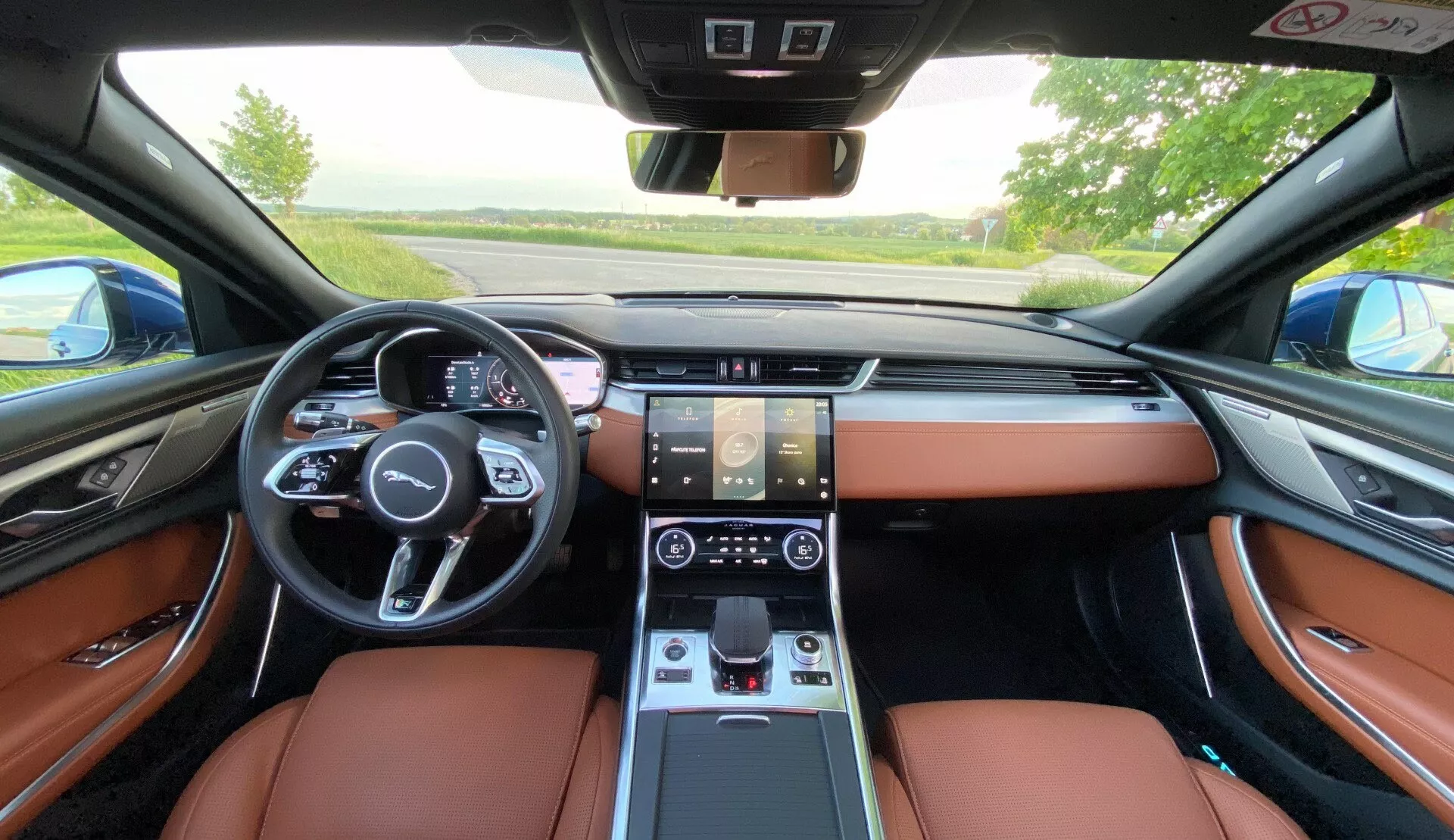 Interior view of 2021 Jaguar XF from backseat perspective, showcasing luxurious brown leather seats, multi-functional steering wheel with controls, digital instrument cluster, dual touchscreen displays for infotainment and climate control, electronic gear shift, and sophisticated dashboard design with integrated climate vents.