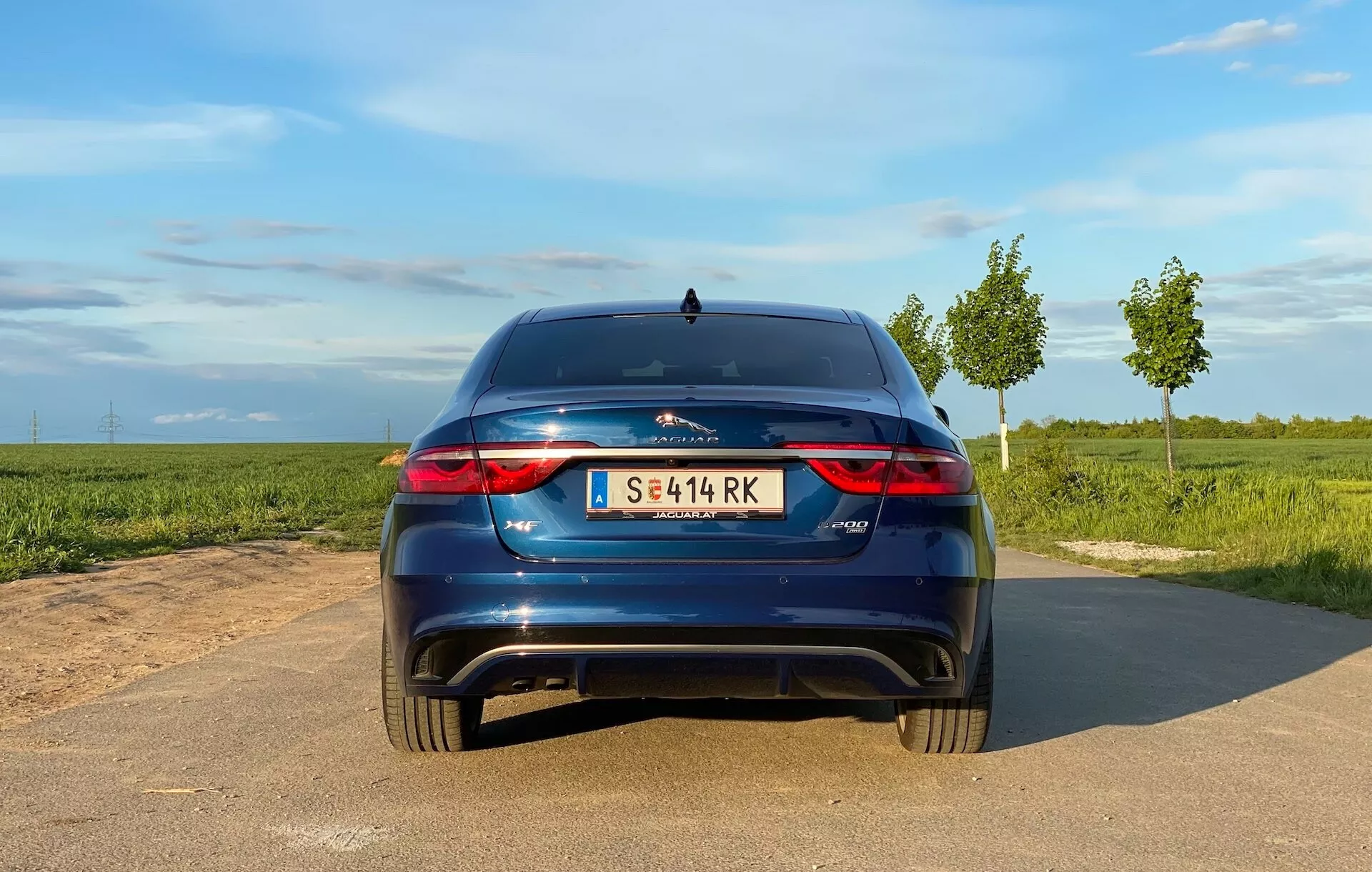 Rear view of a 2021 Jaguar XF hatchback car on a narrow paved road, daylight with clear skies, open fields on either side. Visible features: horizontal LED taillights, dual exhaust outlets, central logo badge, model emblem to the right, central license plate, modest rear spoiler, reflectors on rear bumper, possible parking sensors/camera, shark-fin antenna.