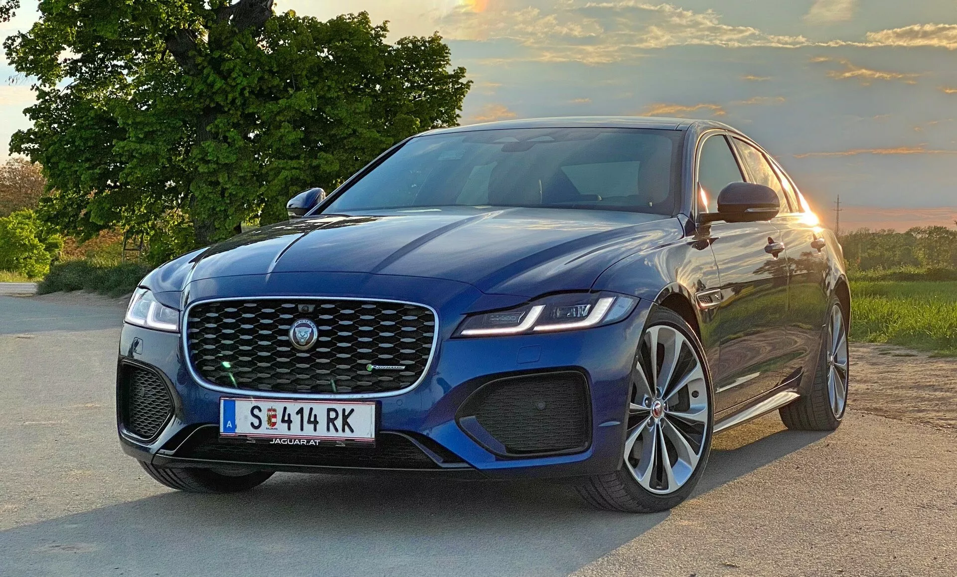 2021 Jaguar XF modern sedan in a three-quarter front view, capturing the large front grille with manufacturer's emblem, sleek angular headlights, sporty front bumper with air intakes, and part of the side profile with raised hood accent lines, large alloy wheels, and side mirrors with integrated indicators. The vehicle is on a road with a natural landscape featuring trees and a sunset sky in the background.