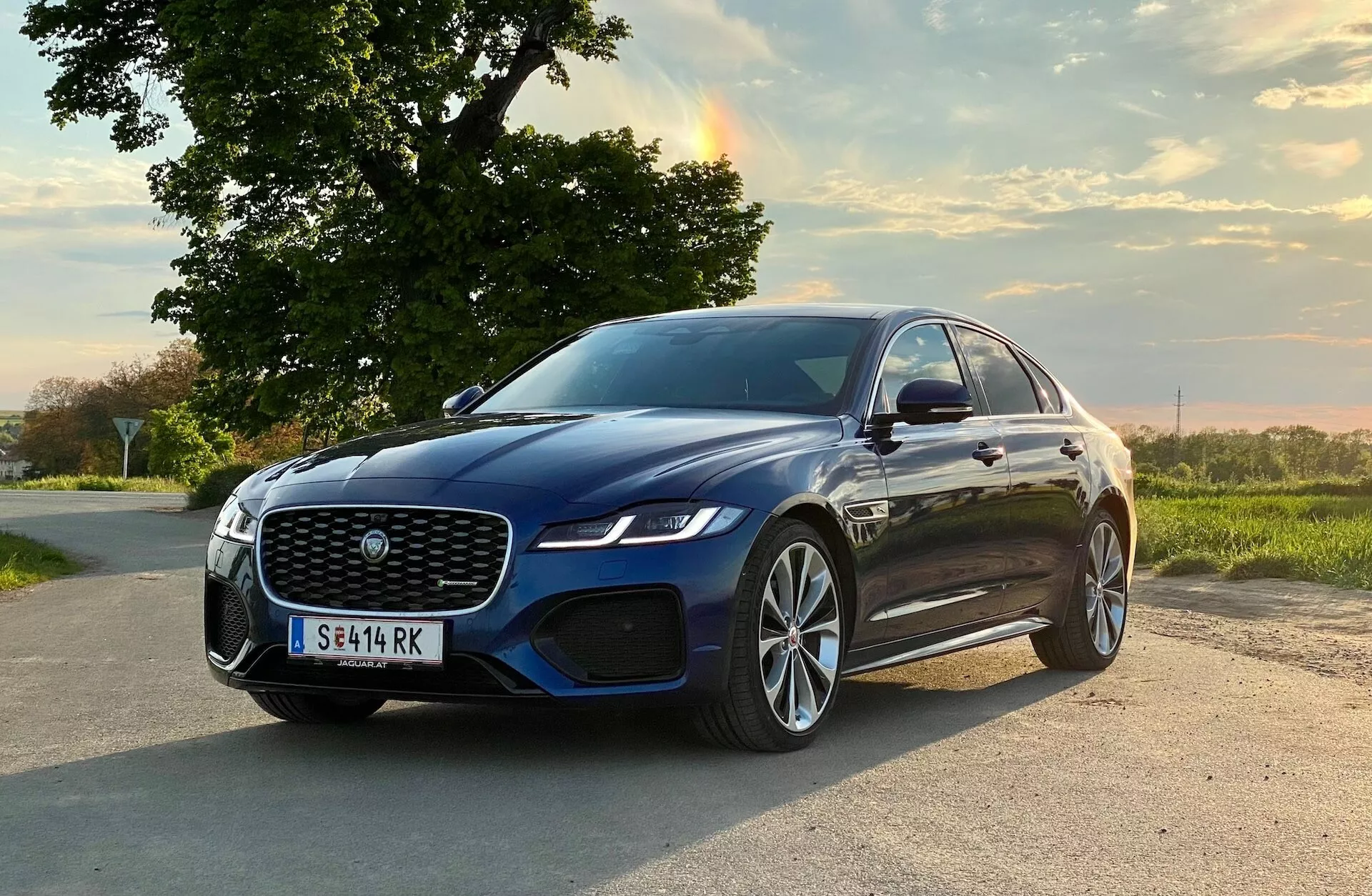 This image depicts the 2021 Jaguar XF, a luxury sedan, positioned diagonally on a rural road. The car faces slightly to the right of the frame, showcasing its front-left quarter including the large mesh grille, sleek LED headlamps, integrated air dam with parking sensors, door-mounted side mirrors potentially with turn signal indicators, multi-spoke alloy wheels with low-profile tires, and aerodynamic body contours with a sleek profile. The scene is set in a serene outdoor environment with warm, natural evening lighting, highlighting the car's premium design and advanced technological features.