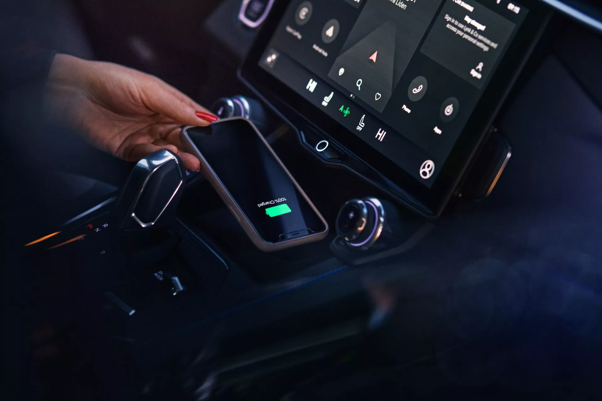 Close-up view of the Lynk & Co 01 2021 center console, showing the vertical infotainment system touchscreen, integrated wireless charging pad with a smartphone charging, ambient LED lighting, and modern gear selector, from the passenger side perspective.