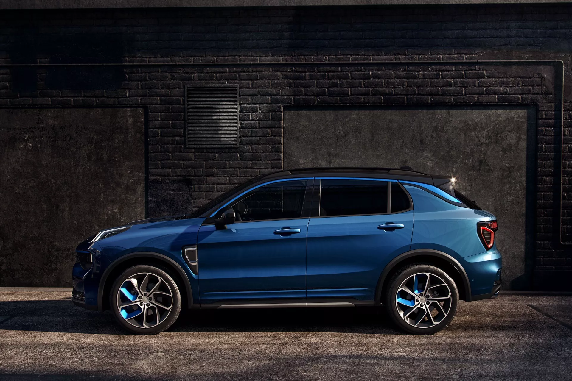 2021 Lynk & Co 01 (1st generation) blue modern SUV in a side view profile highlighting its sporty alloy wheels, dynamic character line, and side profile aerodynamics against an industrial dark brick wall
