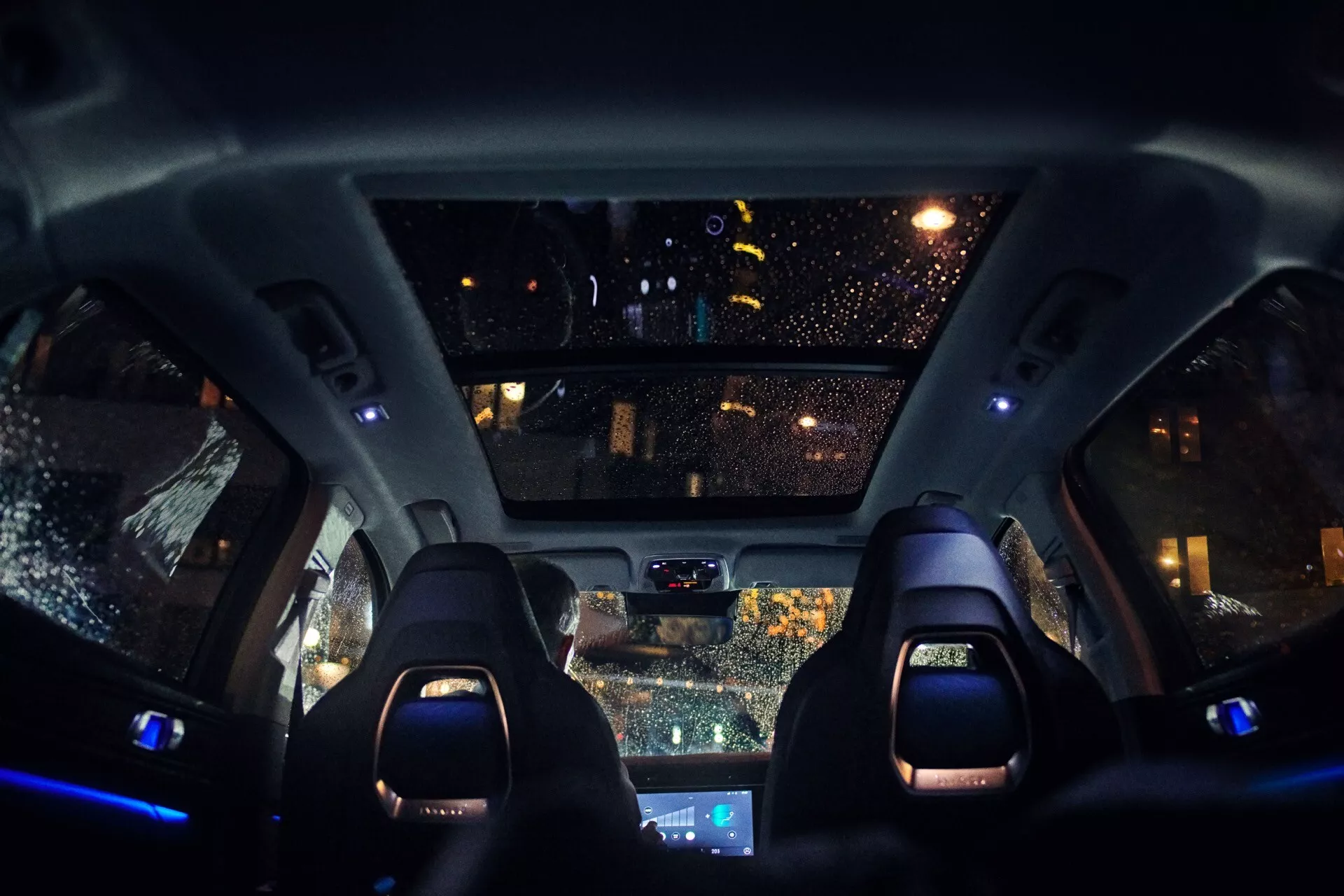 2021 Lynk & Co 01 (1st Generation) interior view at night showcasing sporty front seats with illuminated headrests, modern center console, auto-dimming rearview mirror with sensors, raindrop-covered windshield reflecting city lights, overhead console with cabin lights and possible sunroof controls, and blue ambient interior lighting