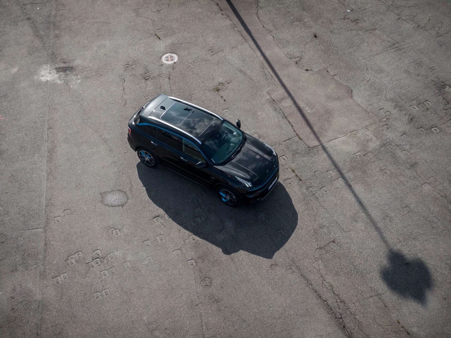 Elevated bird's-eye view of a Lynk & Co 01 2021 model, sleek black sedan with sunroof, positioned diagonally on a paved surface showcasing the entire top side, including roof and hood, in early morning or late afternoon sunlight casting a long shadow