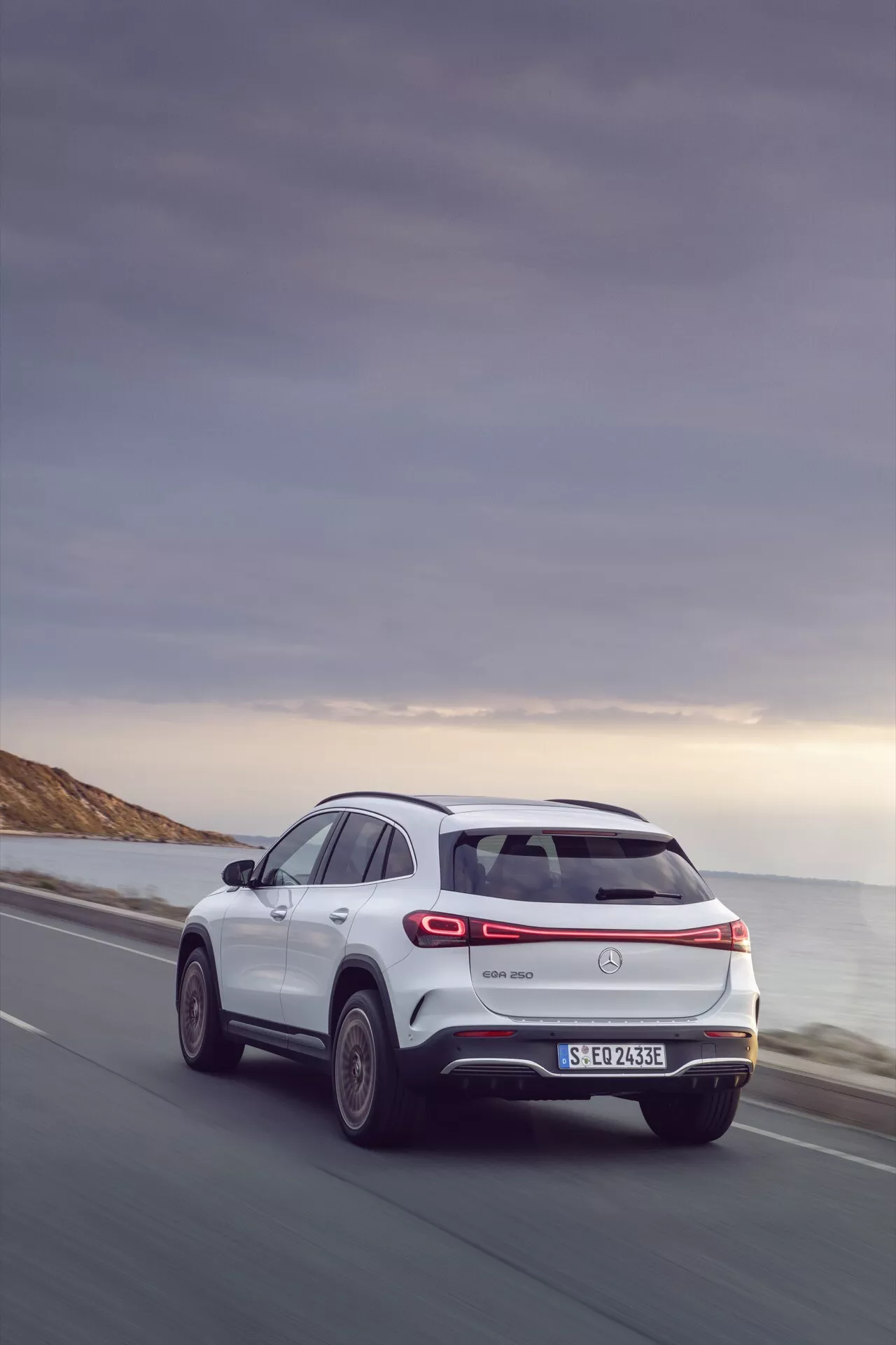 2021 Mercedes-Benz EQA 243 Rear Three-Quarter View on Coastal Road, Highlighting Side Profile, Rear Design with LED Taillights, Manufacturer's Logo, Rear Wiper, Bumper with Integrated Reflectors and Parking Sensors, Decorative Exhaust and Electric Model Indications, Big Wheels with Multi-Spoke Alloy Design, Indicating an SUV with Potential Advanced Aerodynamics and All-Wheel-Drive Capabilities under Dusky Sky