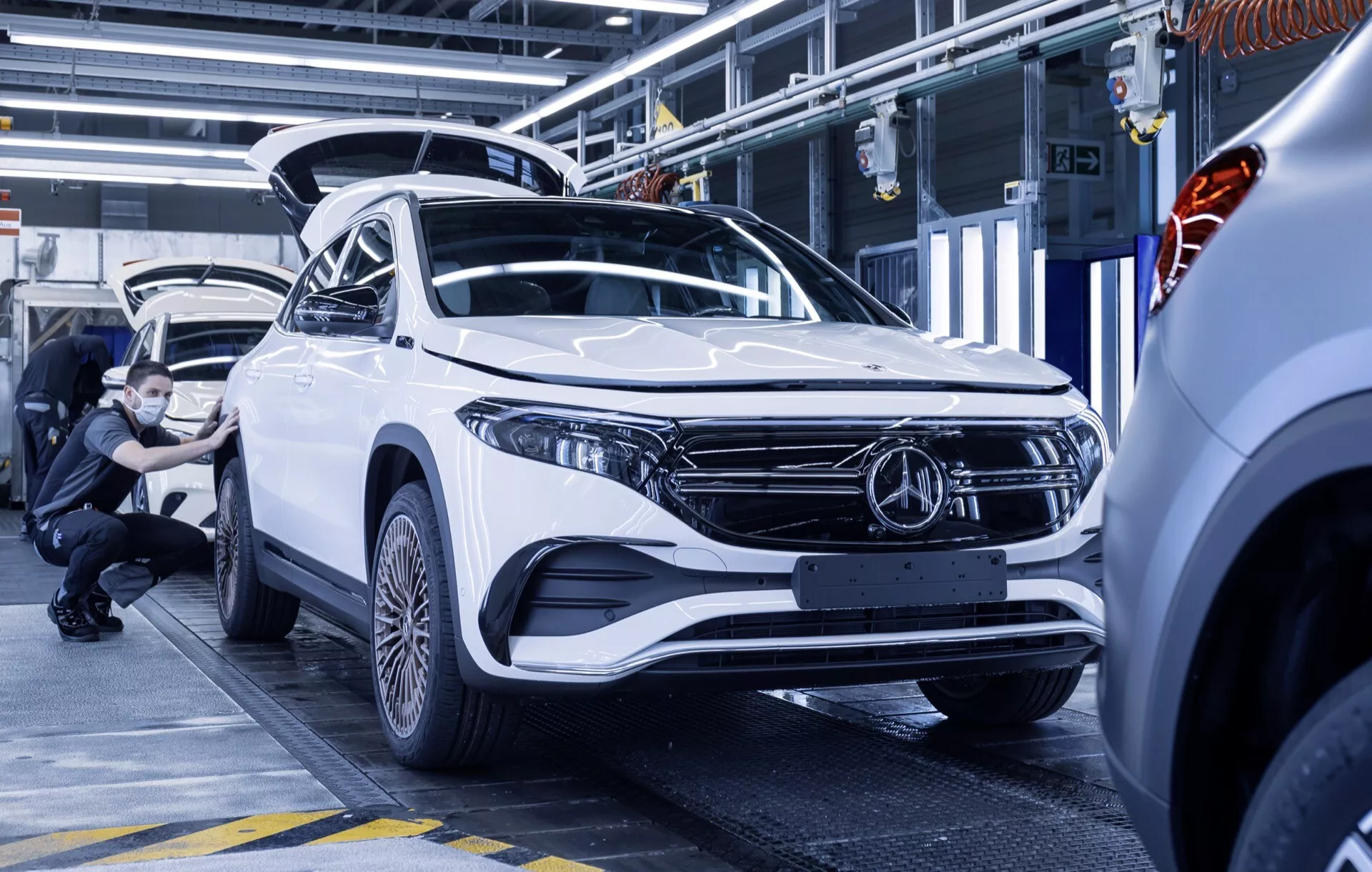 2021 Mercedes-Benz EQA in white, showcasing a modern SUV design with an angular front three-quarter view inside a car assembly or inspection facility, highlighting the vehicle's sleek LED headlights, aerodynamic front bumper, and prominent grille badge.