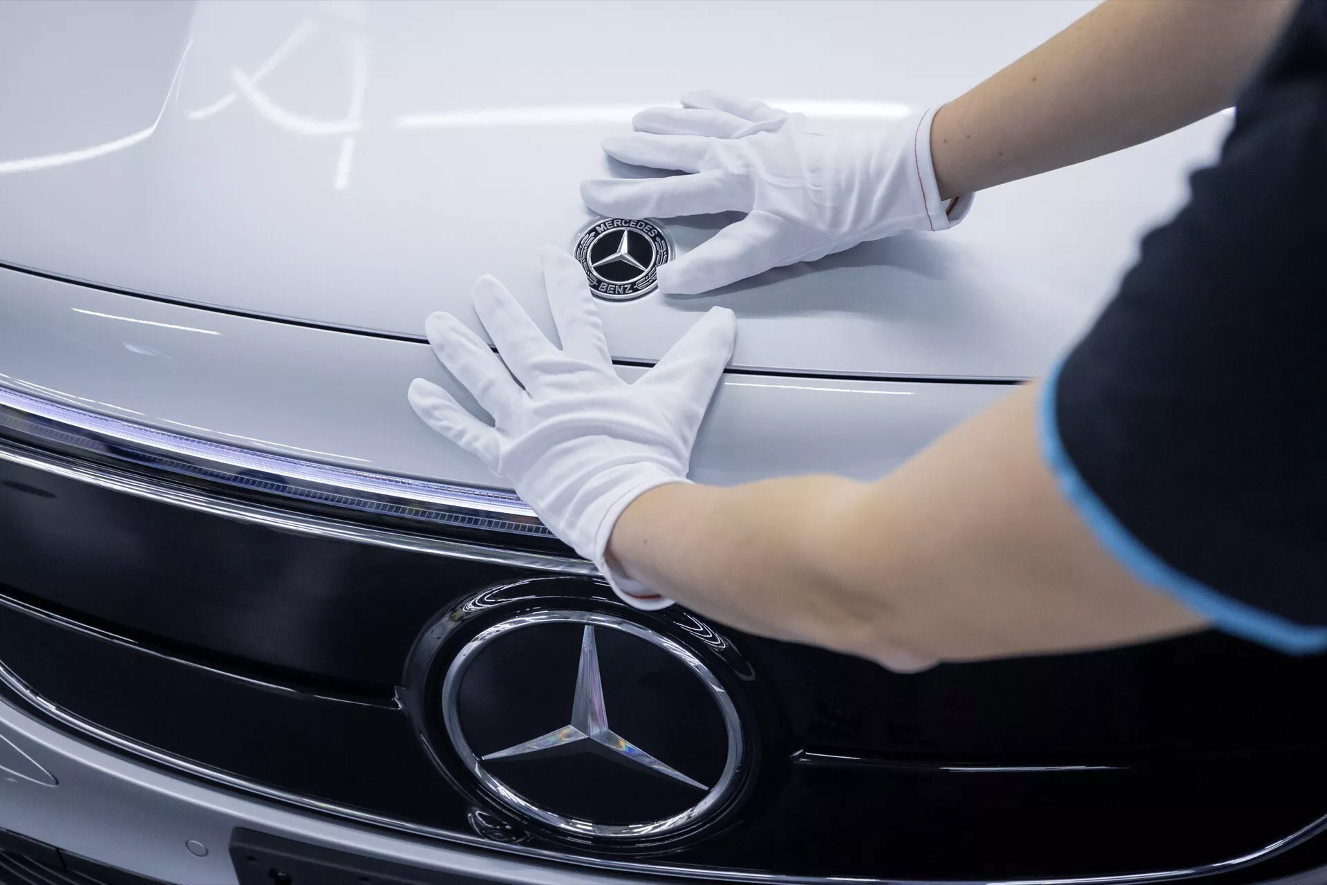 Close-up view of the Mercedes-Benz EQA 2021's grille and emblem with white-gloved hands touching, showcasing chrome detailing and LED headlight technology.