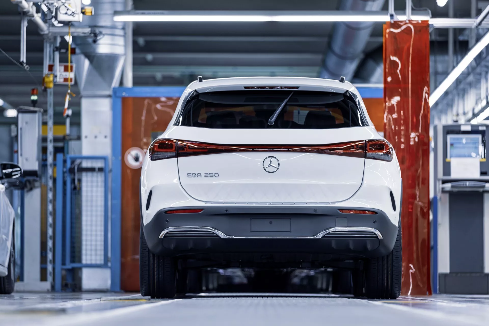Rear view of a white Mercedes-Benz EQA 250 electric vehicle in a modern industrial setting, highlighting the tail lights, rear bumper, and Mercedes branding with a clear focus on design and technological features.