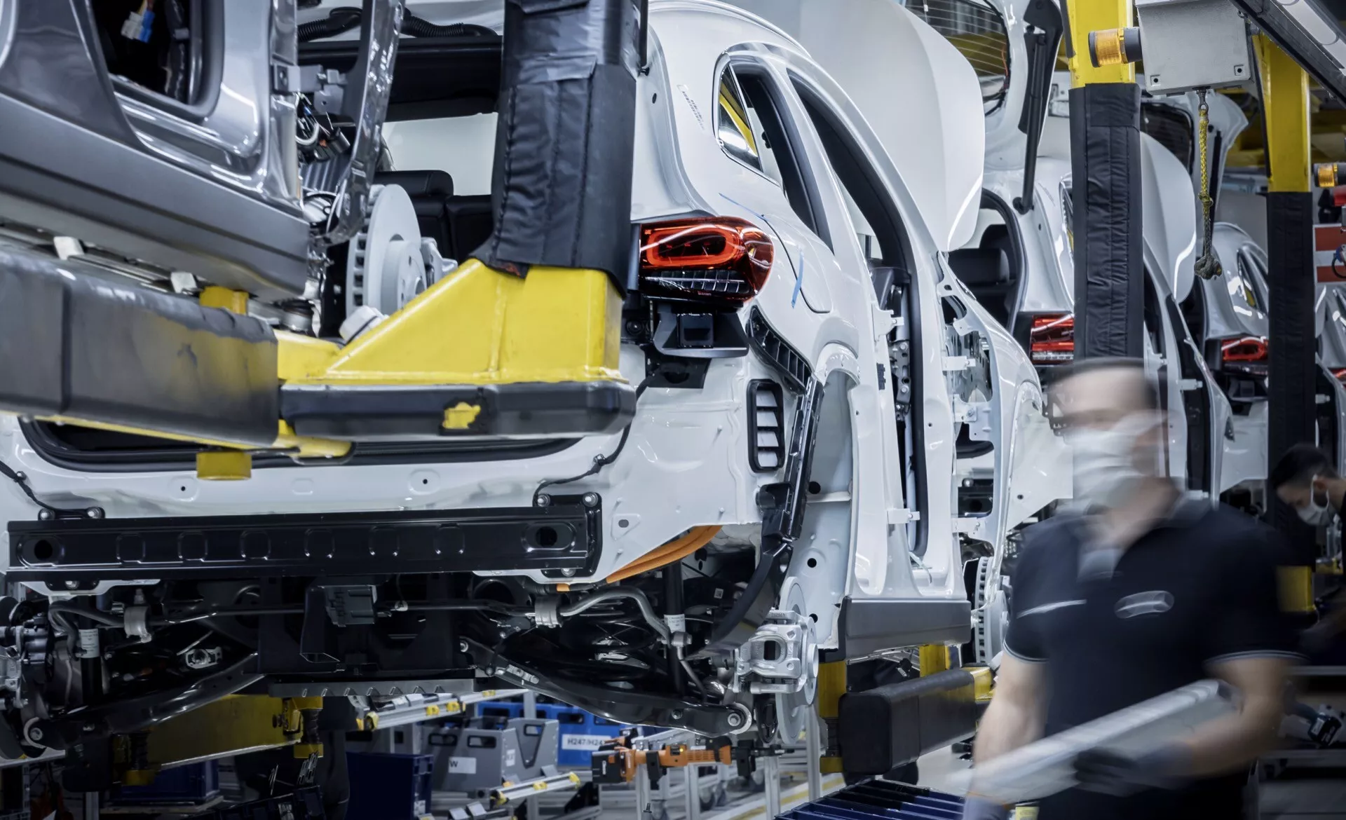 Early stage assembly of Mercedes-Benz EQA 2021 on factory line, showing car's body shell and unibody chassis supported by automated robotic arms with visible rear structure and assembly workers in background