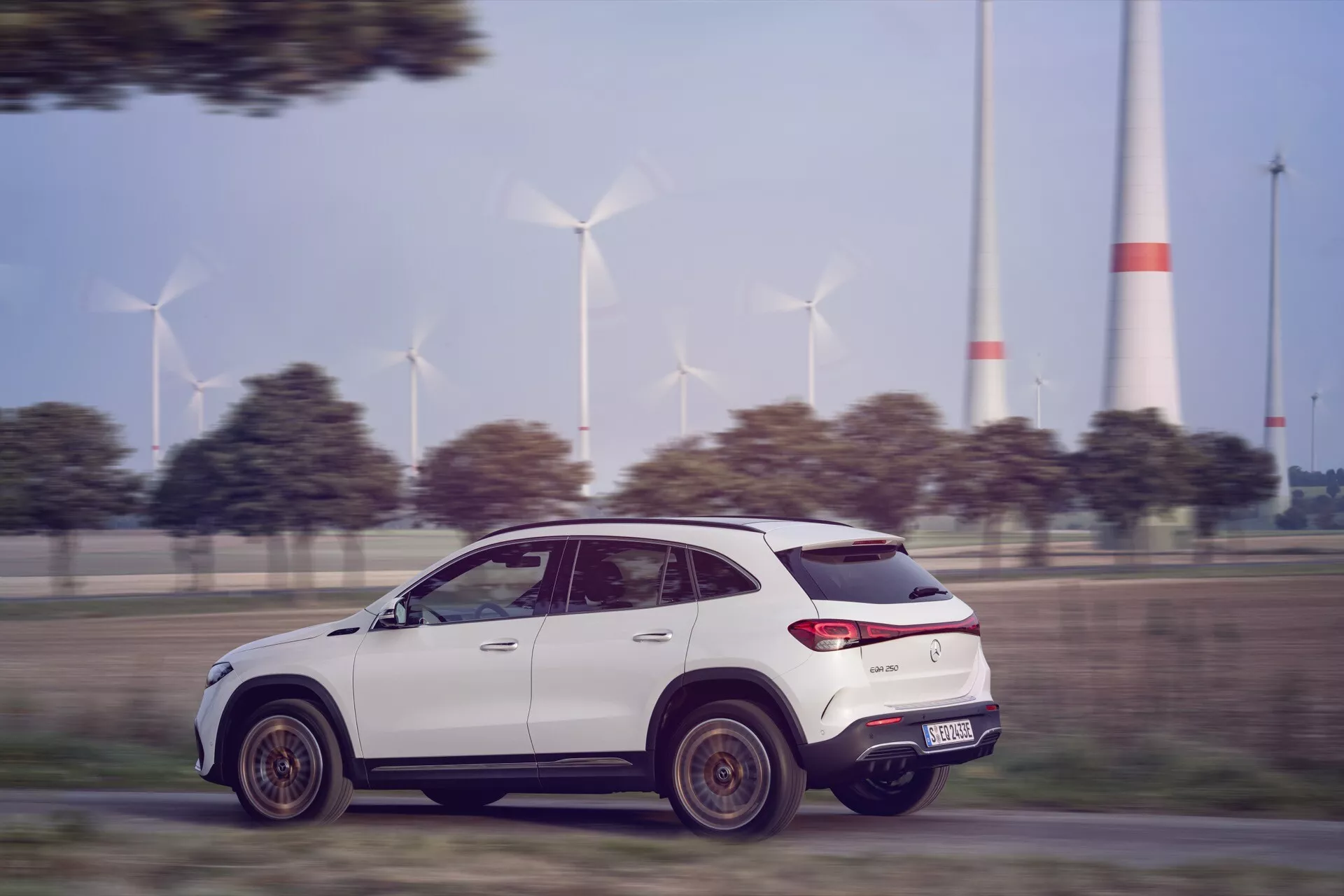 2021 Mercedes-Benz EQA in dynamic motion, side view profile of a modern SUV on a paved road with wind turbines in the background, showcasing aerodynamic design, LED tail lights, and high ground clearance