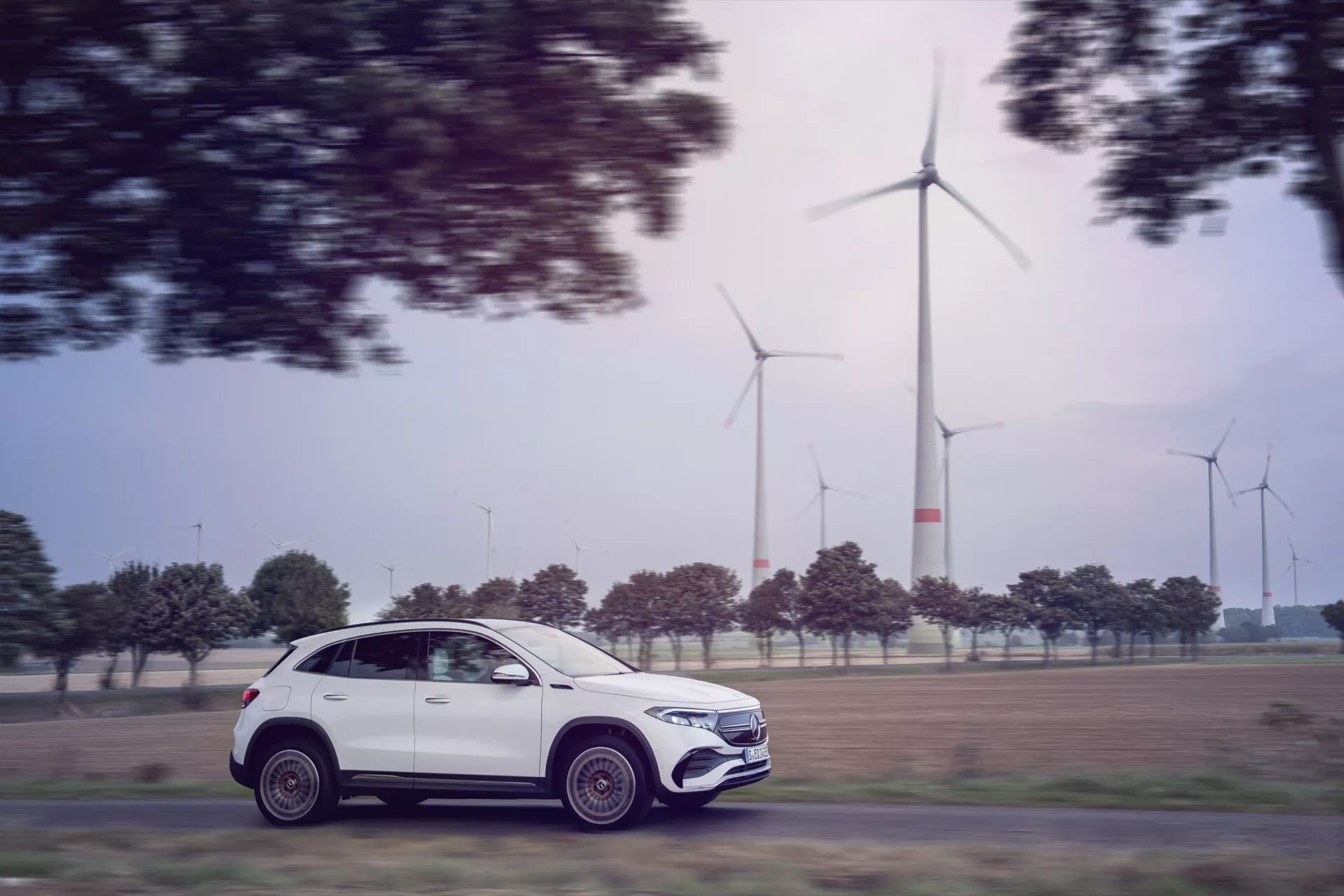 2021 Mercedes-Benz EQA 243 in motion captured with panning photography technique, showcasing its modern SUV profile, intricate alloy wheels, and eco-friendly design against a background of wind turbines