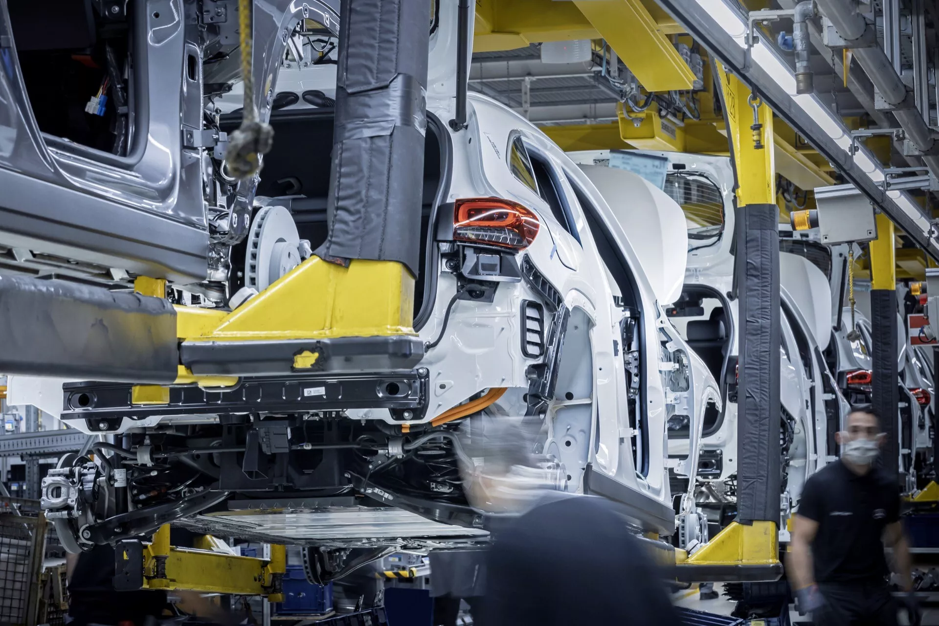 2021 Mercedes-Benz EQA side profile assembly in progress inside manufacturing plant, featuring rear half suspended by yellow robot arm, visible unibody structure, and partial interior components.