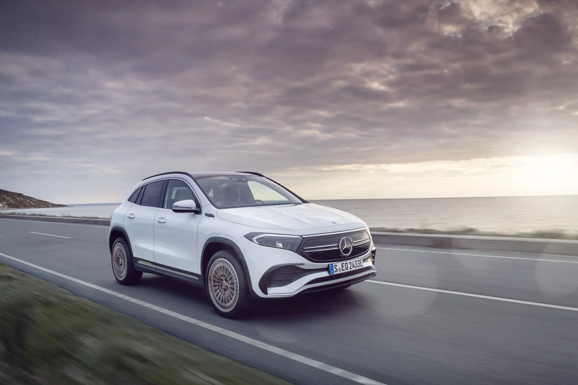 2021 Mercedes-Benz EQA in white, showcasing its dynamic movement and luxurious design on a scenic coastal road, highlighting its LED headlights, Mercedes emblem, and aerodynamic silhouette at sunset.