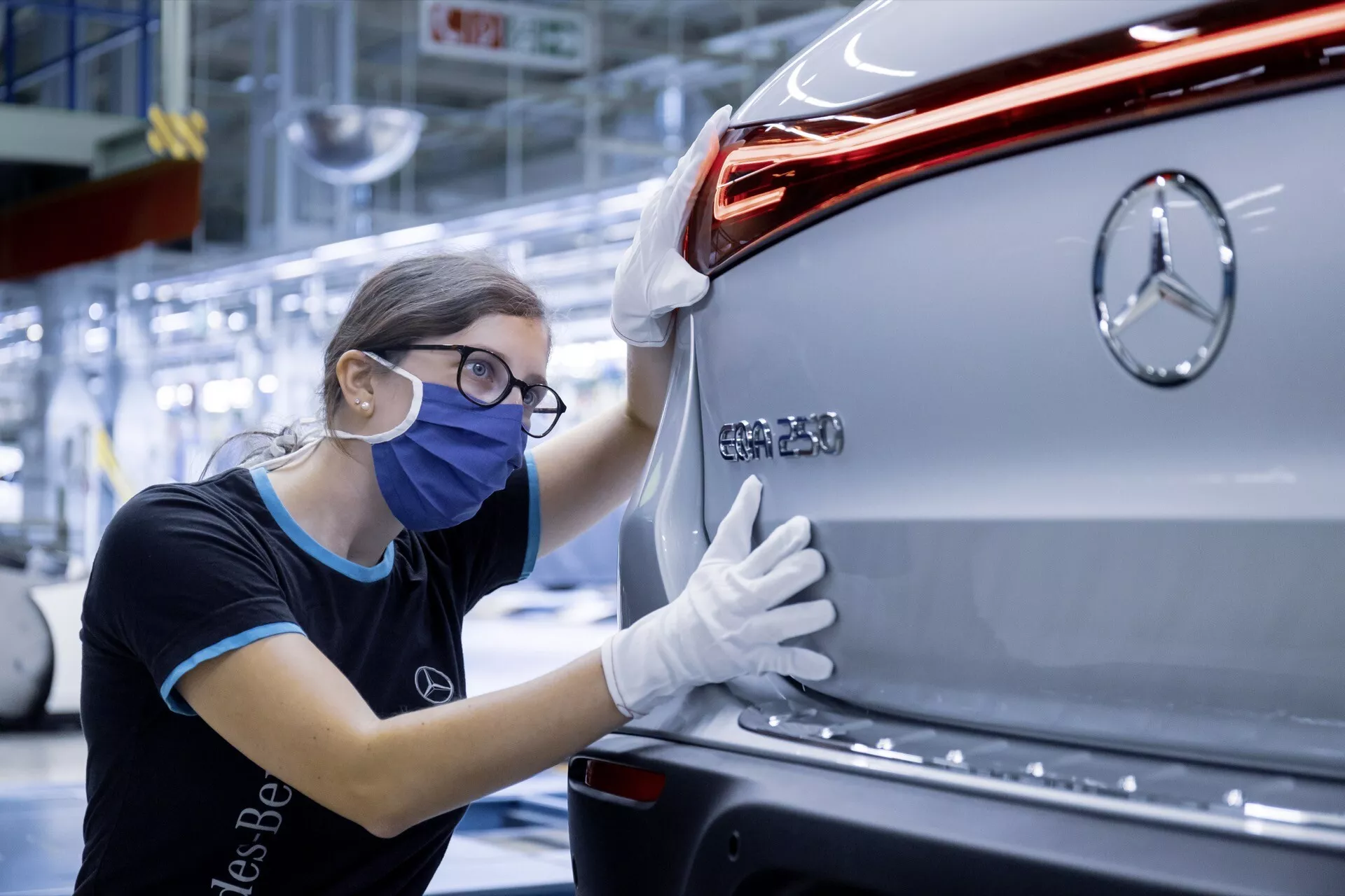 Rear view of a silver Mercedes-Benz EQC 2021 showcasing the brand badge, modern LED taillight, sleek car design, and rear bumper, with a quality control worker inspecting the vehicle in a factory setting