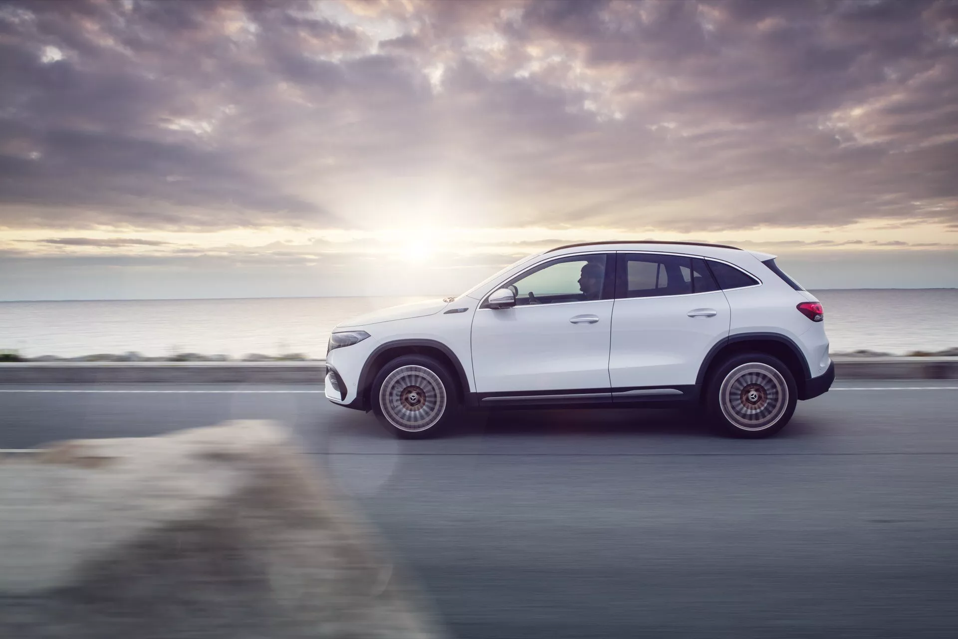 Dynamic side profile of 2021 Mercedes-Benz EQA SUV in white, showcasing its sleek design and rugged stance on a beachside road, highlighted by sunset lens flare and technology features like alloy wheels and possible LED lighting.