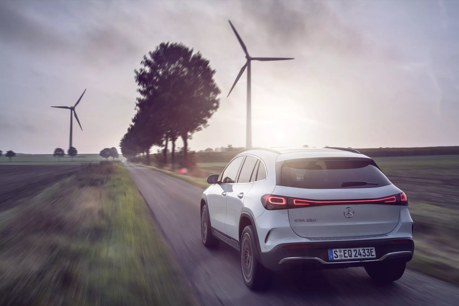 2021 Mercedes-Benz EQA electric SUV in motion, rear three-quarter view showcasing aerodynamic design and modern technology, with landscape of renewable energy wind turbines in the background