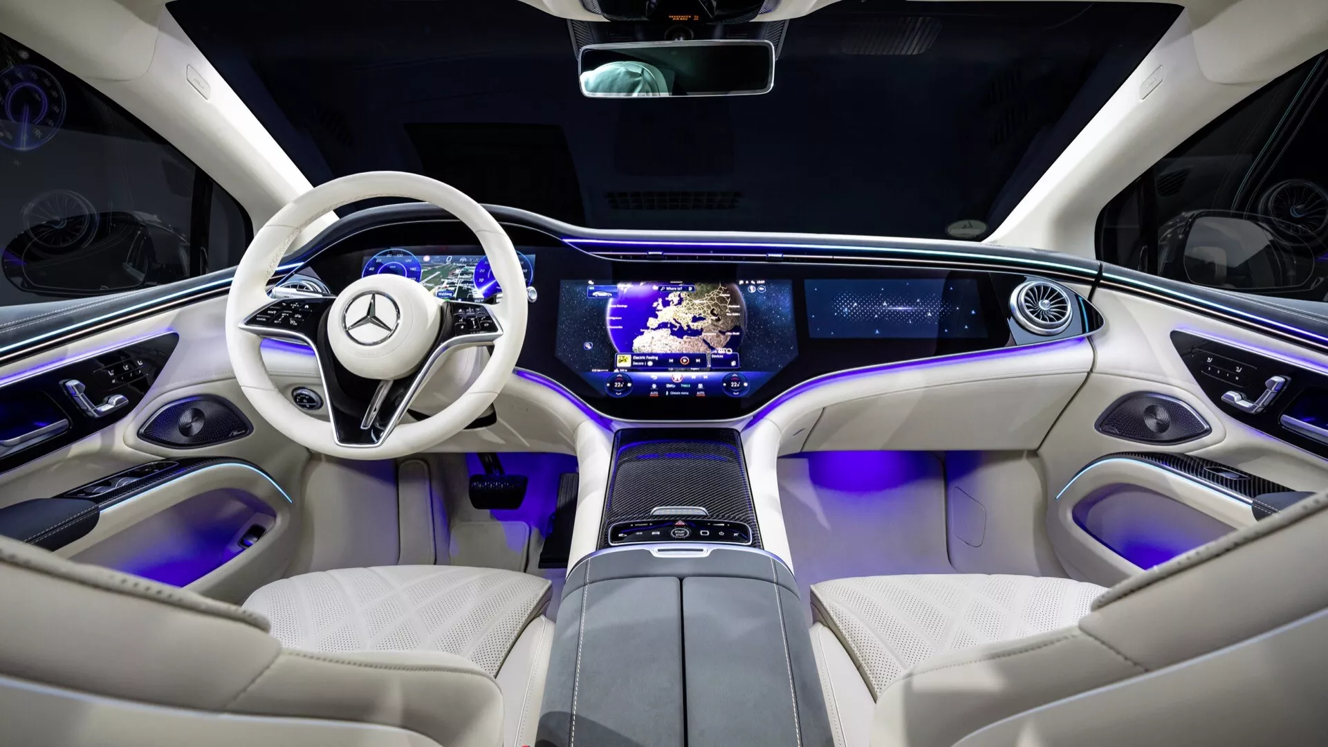 2024 Mercedes-Benz EQS facelift interior from rear seat perspective highlighting technology features and luxury elements, including a two-spoke luxury steering wheel with controls, sleek dashboard with dual-screen setup, center console with touch-sensitive controls, blue ambient lighting, cream colored leather seats, and panoramic sunroof