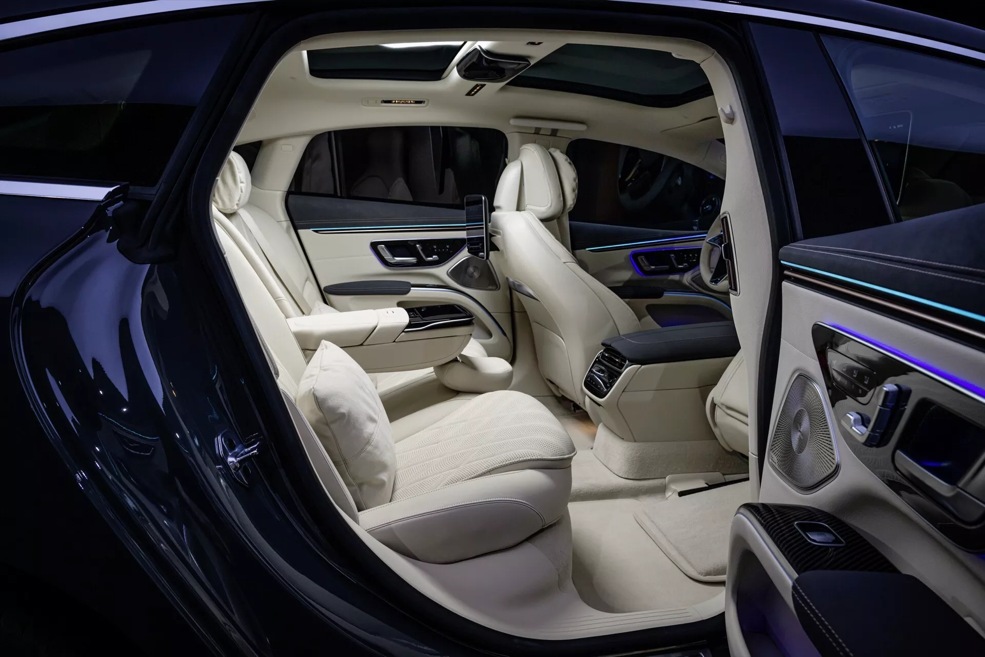 2024 Mercedes-Benz EQS 297 facelift interior showcase including sophisticated light-colored leather seats, sleek dashboard design with digital display, high-end door panels with controls, extended central console, rear passenger entertainment system, premium carpet flooring, and ambient cabin lighting from an exterior side view with the front passenger door open.