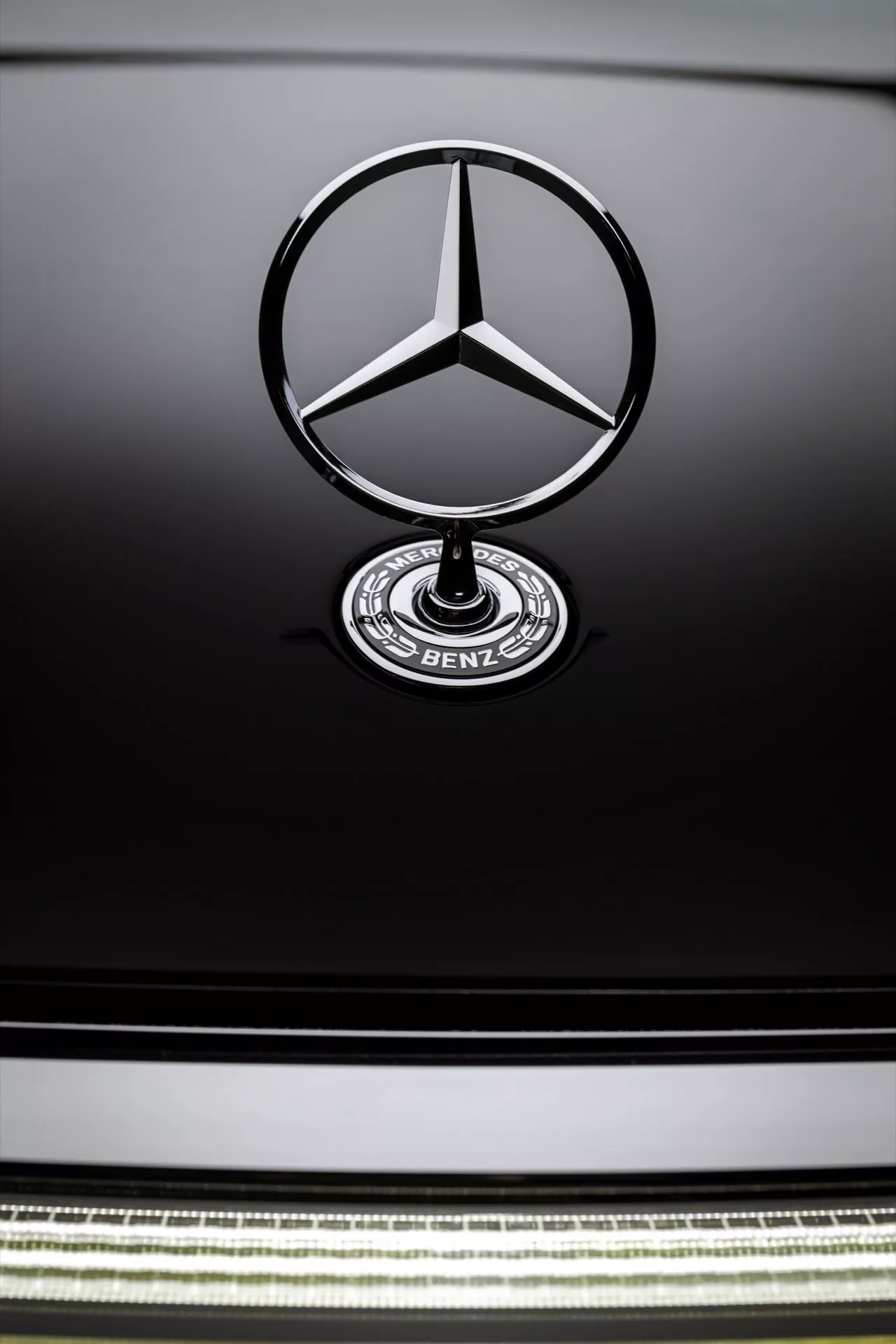 Close-up view of the Mercedes-Benz EQS 2024 facelift emblem and grille, featuring the signature three-pointed star and ‘BENZ’ badge on a dark-colored hood, with a detailed chrome strip in the grille suggesting advanced aerodynamics.