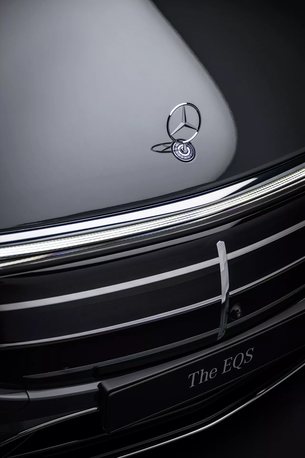 Close-up view of 2024 Mercedes-Benz EQS front grille and badge, showcasing chrome emblem against a dark contoured hood, with elegant horizontal chrome slats and 'The EQS' inscription indicating luxury electric vehicle design