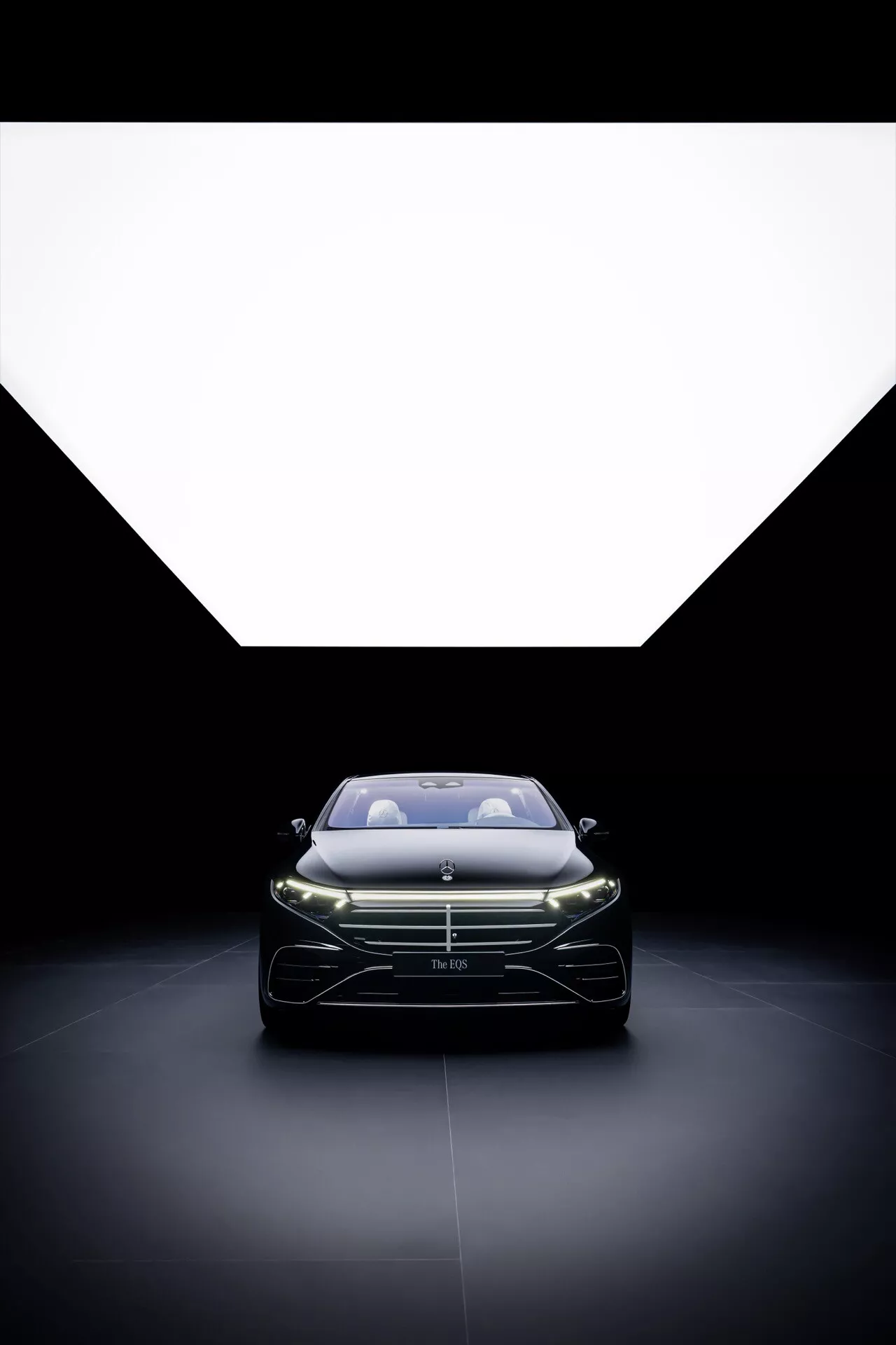 2024 Mercedes-Benz EQS Facelift featuring forward-facing view with illuminated LED headlights and pronounced front grille, captured from a low angle to highlight sleek design and aerodynamic contours against a dramatically lit background.