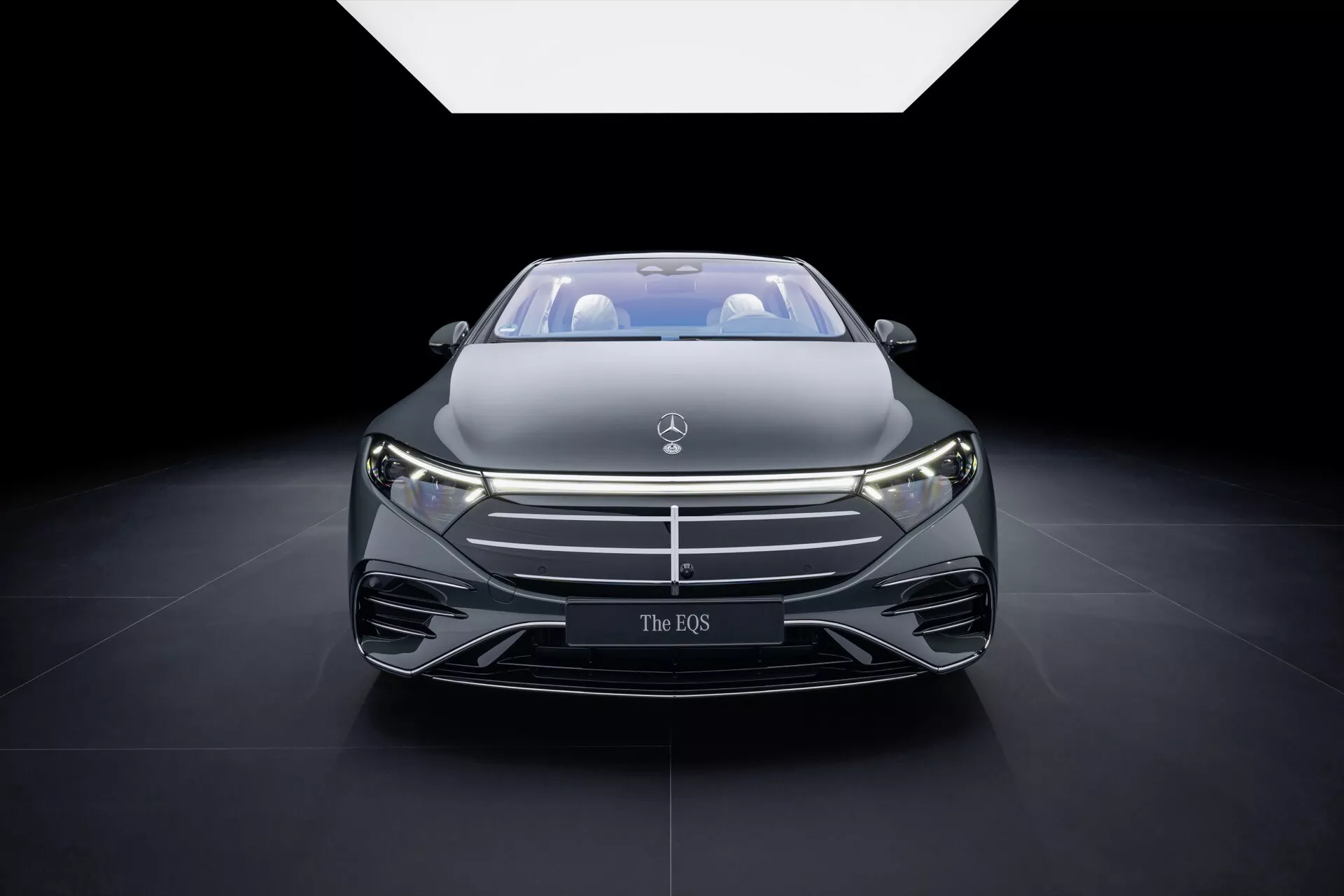Front view of the Mercedes-Benz EQS 2024 (297-Facelift) featuring prominent grille with badge, sleek LED headlights, and aerodynamic hood design, displayed under dramatic spotlight at an auto show