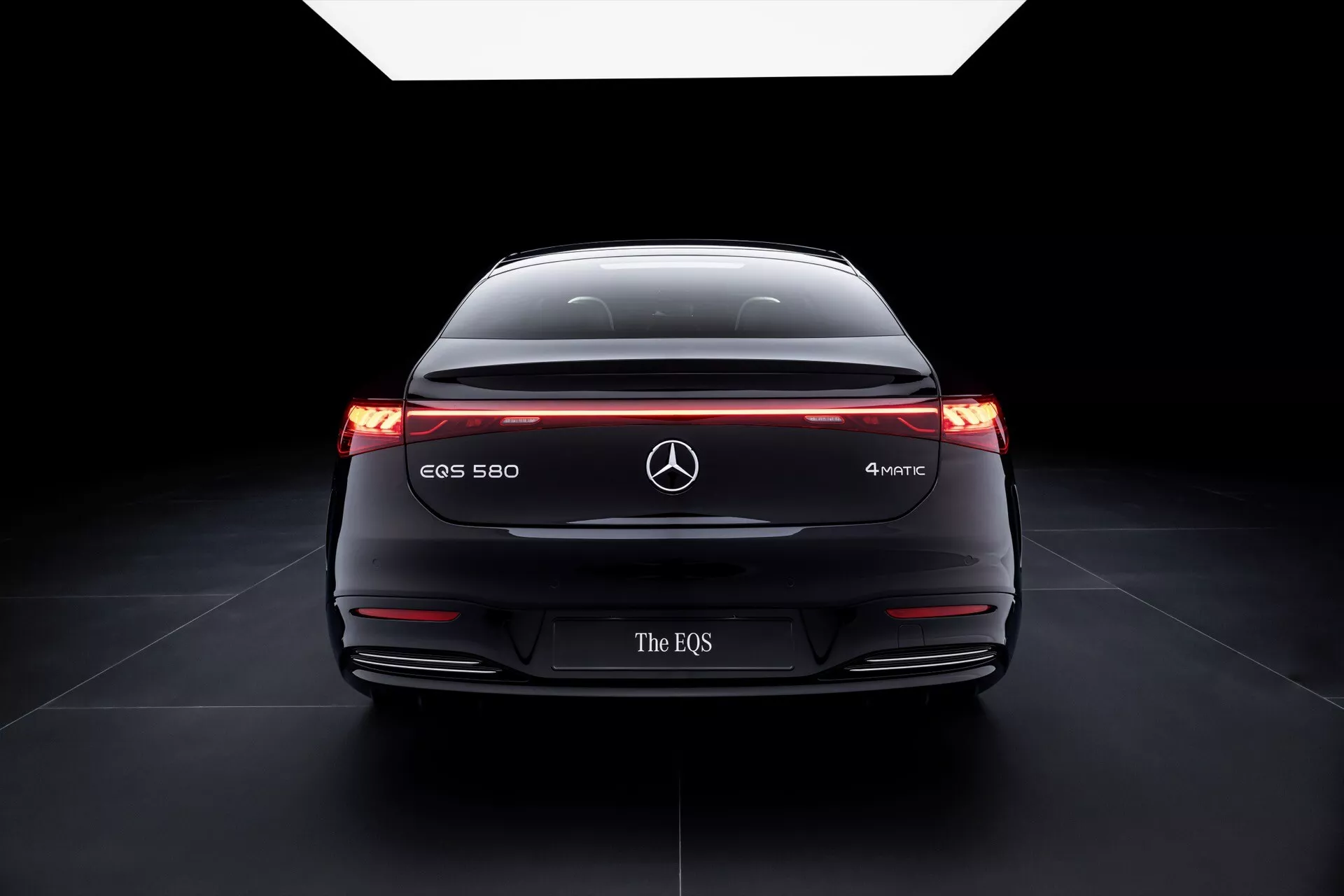 Rear view of 2024 Mercedes-Benz EQS 580 4MATIC showcasing its wide stance, illuminated taillights with horizontal light band, model designation and advanced safety technologies against a dark gradient background emphasizing its aerodynamics and electric vehicle design.