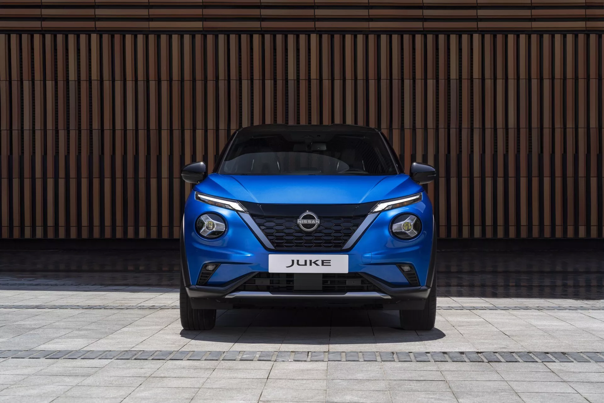 Front view of a 2019 Nissan Juke F16 in blue, showcasing sleek LED headlights, V-motion grille, and sporty bumper against a contrasting horizontal-lined wall background