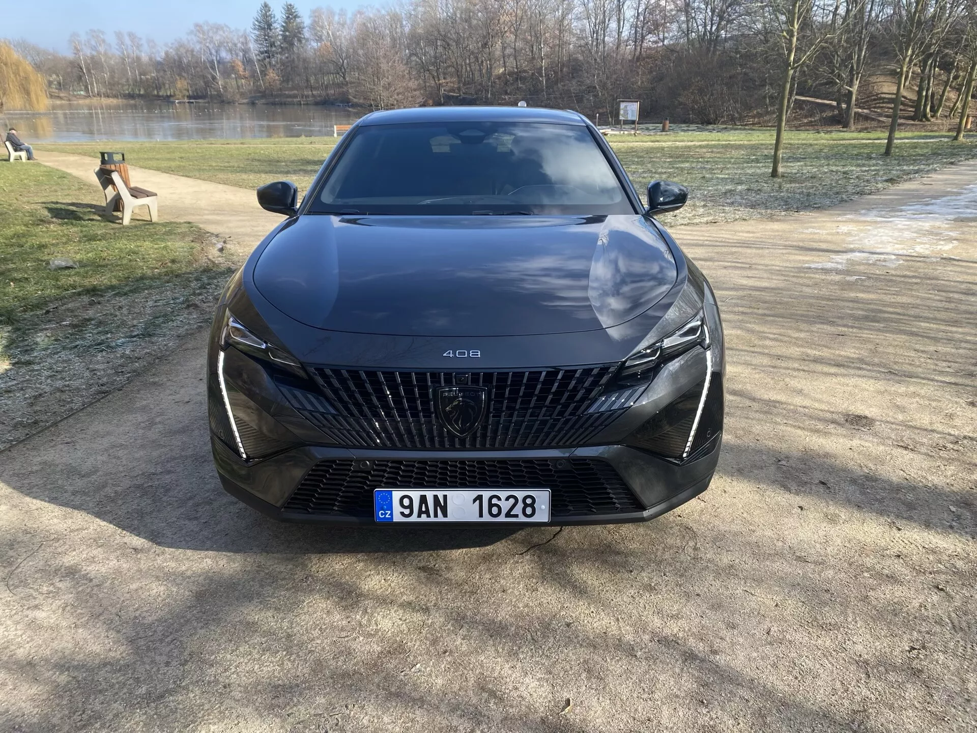 2023 Peugeot 408 front view parked on a gravel surface with trees and water backdrop, highlighting its large grille with diamond-shaped patterns, sleek LED headlights, sculpted hood lines, and advanced driver assistance features