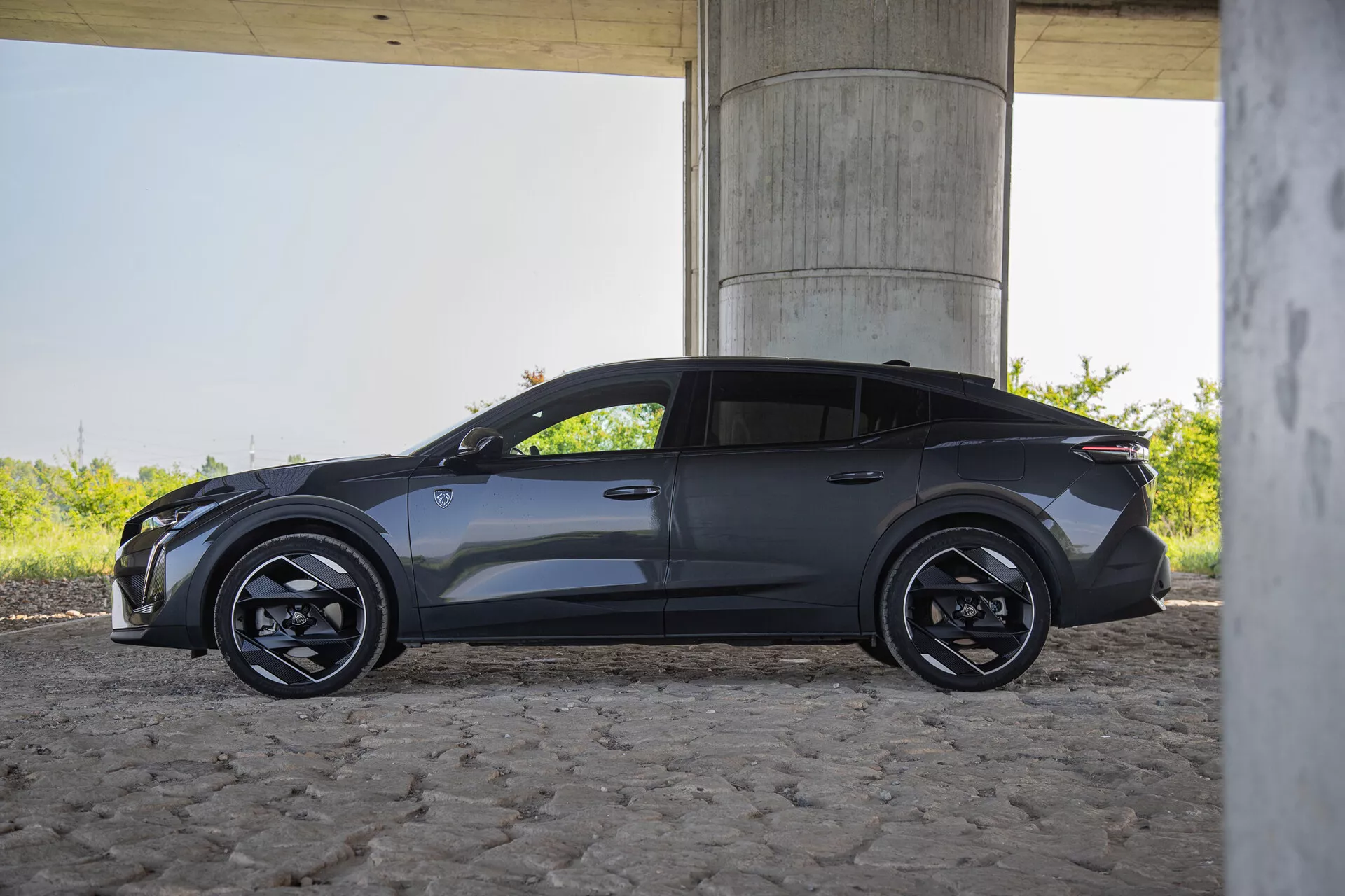 2023 Peugeot 408 side profile outdoor under bridge, featuring sleek design, electric/hybrid closed grille, angular LED headlights, multi-spoke black wheels, low-profile tires, aerodynamic side mirrors with integrated signals, recessed door handles, dynamic character line, sporty rear with compact spoiler, metallic paint in natural daylight
