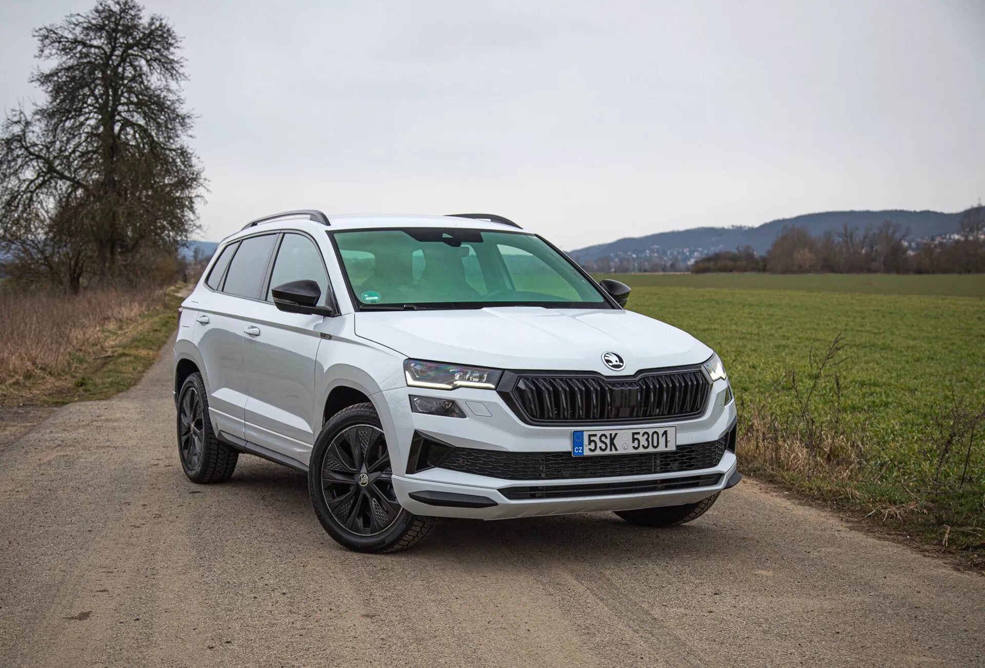 2021 Škoda Karoq white SUV parked on a country dirt road, showcasing front-left quarter view including grille, headlights, and wheel design, with a focus on modern features like LED lighting and parking sensors.