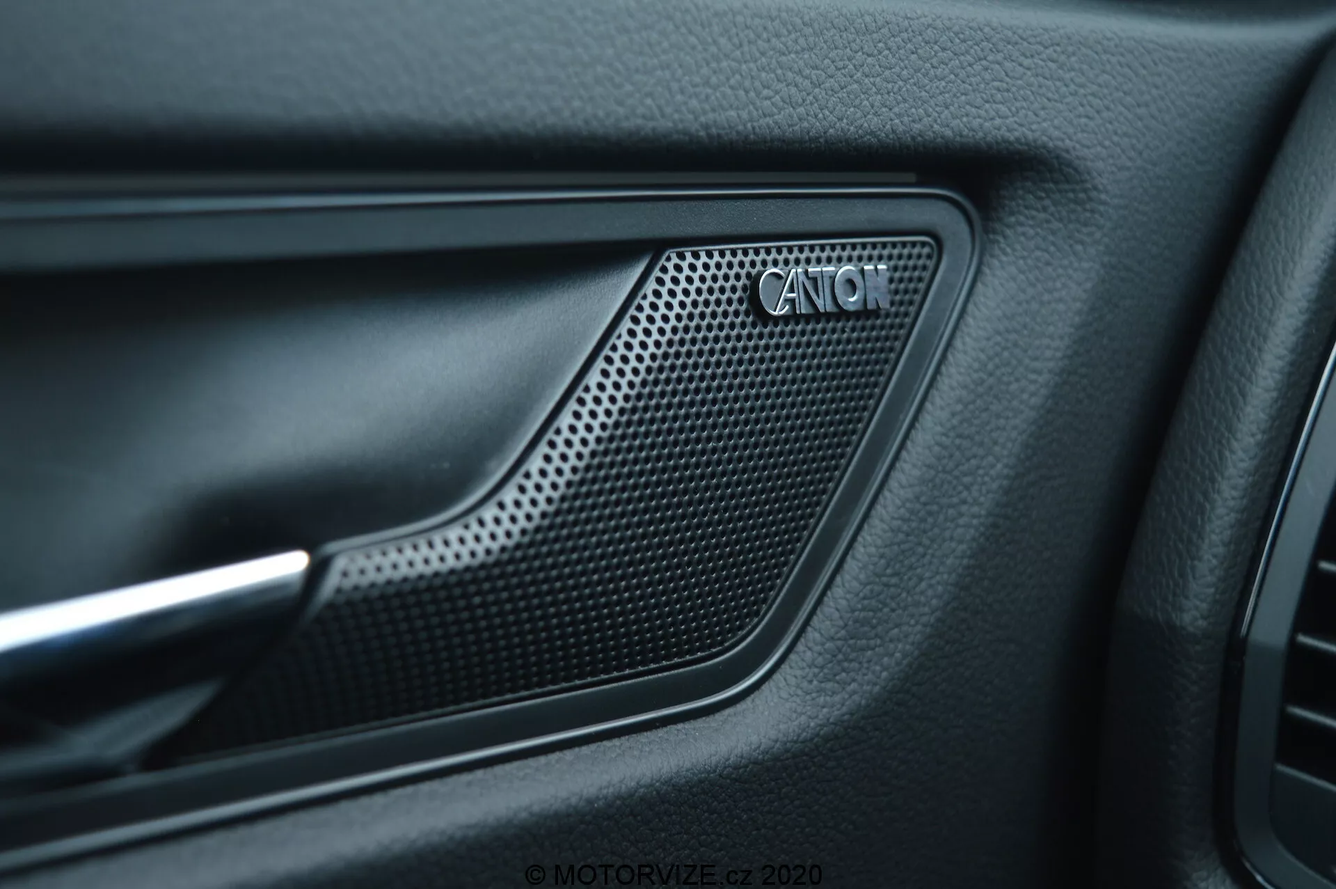 Close-up view of the 2017 Skoda Kodiaq car interior door panel showcasing the CANTON speaker grille. The grille, made of a perforated, honeycomb-like pattern material, is positioned within a recessed area of the door panel framed by an indented outline. Matte black, textured material surrounds the grille, alongside a contrasting metallic accent, reflecting the sophisticated interior design and premium audio system.