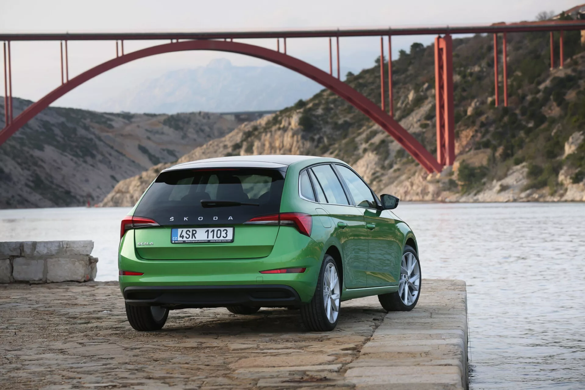 2019 green Skoda Scala parked by water with a rear and side profile view, showcasing taillights, rear hatch, logo, and model designation by a red bridge and mountains backdrop