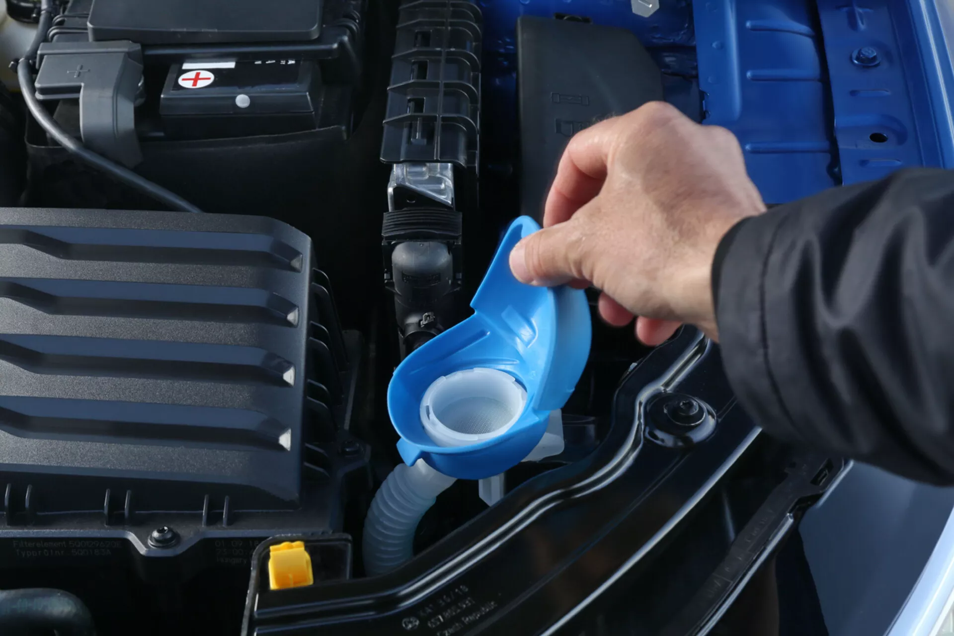 Close-up view of Skoda Scala 2019 engine bay highlighting the refill of windshield washer fluid using a blue funnel, showcasing the battery, engine cover, and car structure