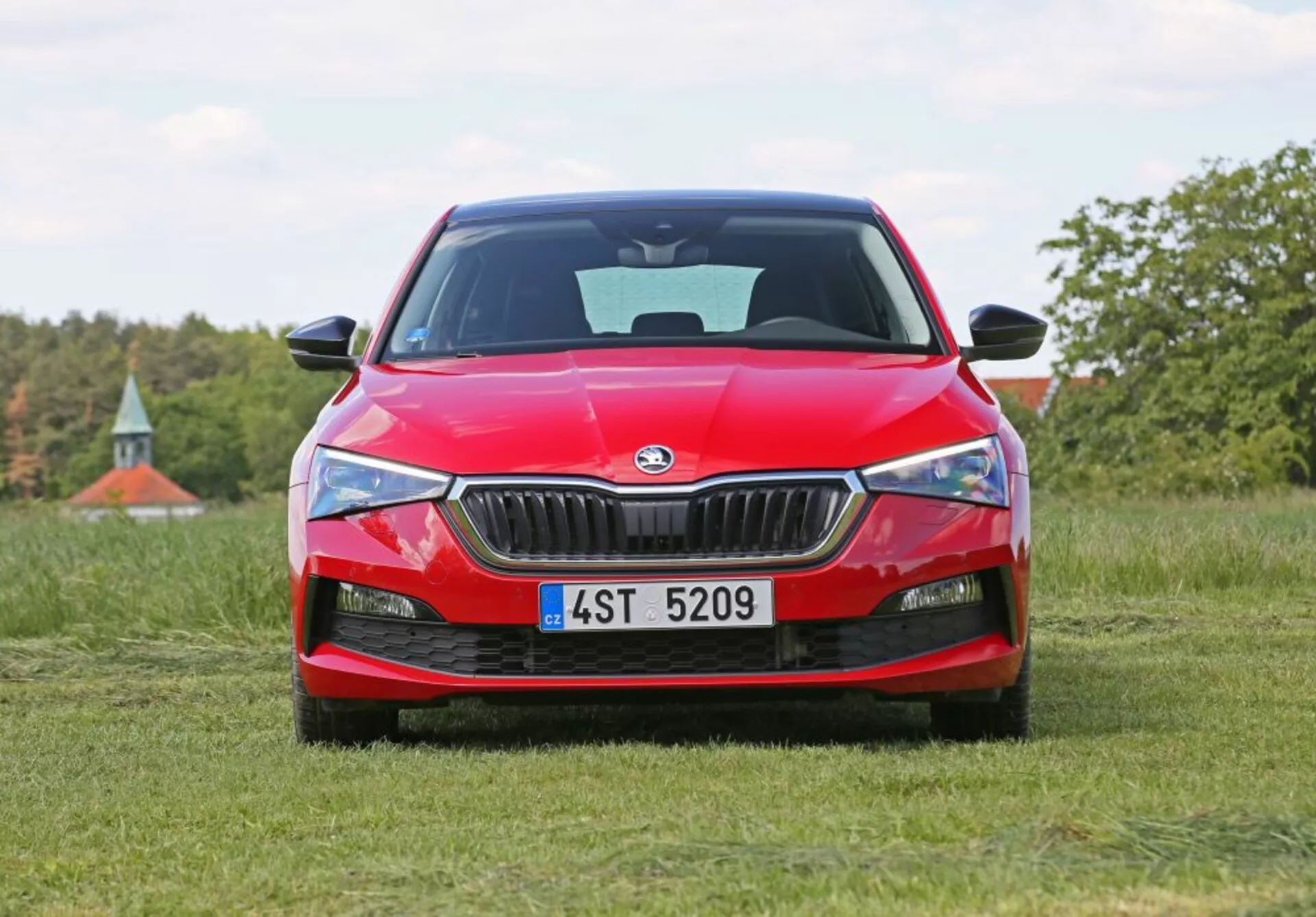 2019 Skoda Scala in bright red, showcasing modern LED headlight technology, angular chrome-trimmed grille, and sporty front fascia design, photographed at a low angle on a grass field with a natural landscape background