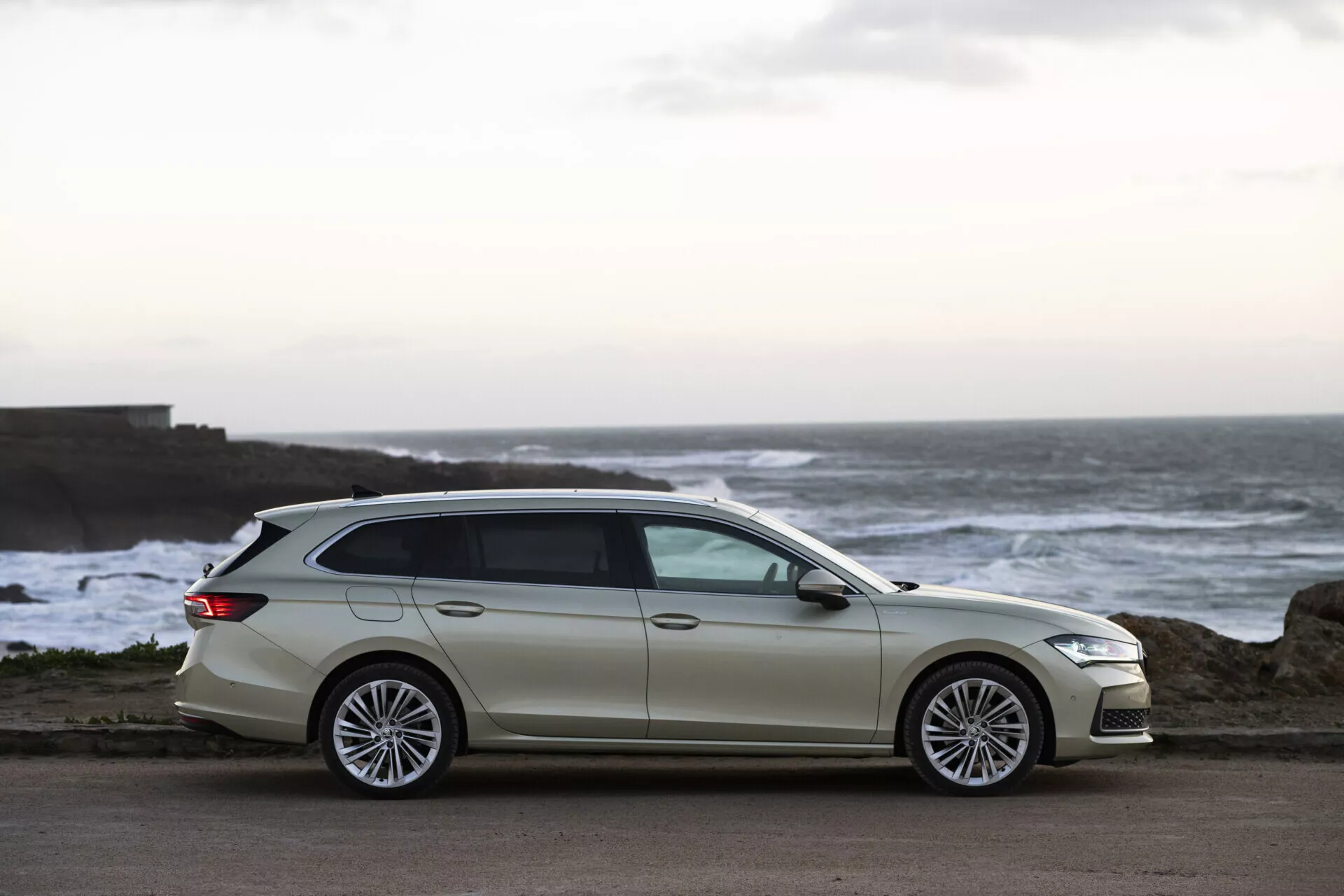 2024 Skoda Superb Combi in a light shade parked near the coast at twilight, highlighting its modern design, streamlined bodywork, and premium multispoke alloy wheels