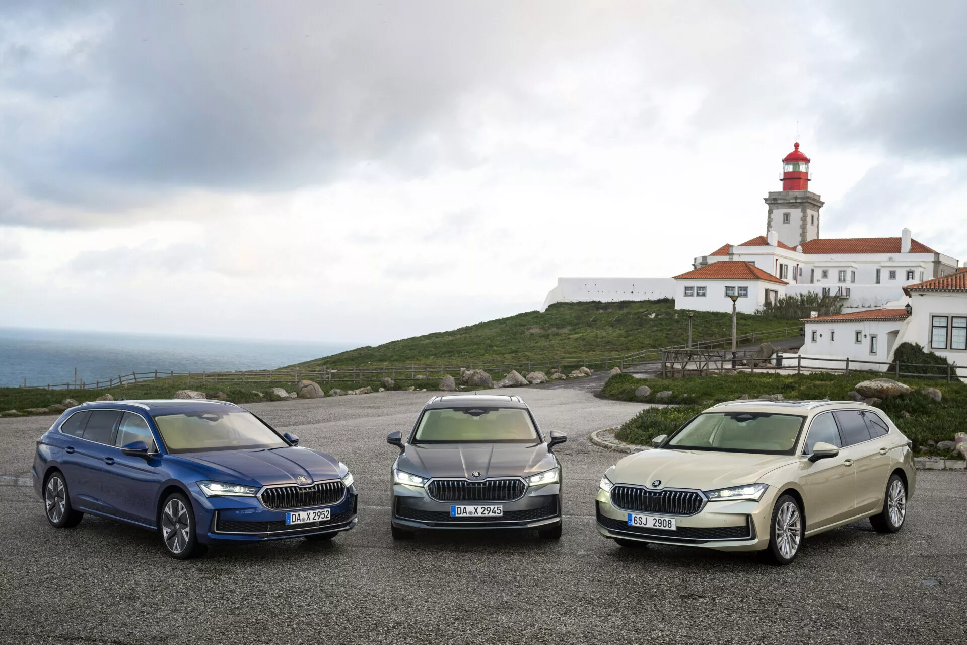 2024 Skoda Superb Combi models in dark blue, metallic grey, and pale gold parked side-by-side showcasing LED headlights, chrome grilles, and alloy wheels with a lighthouse and building structure in the background under a cloudy sky