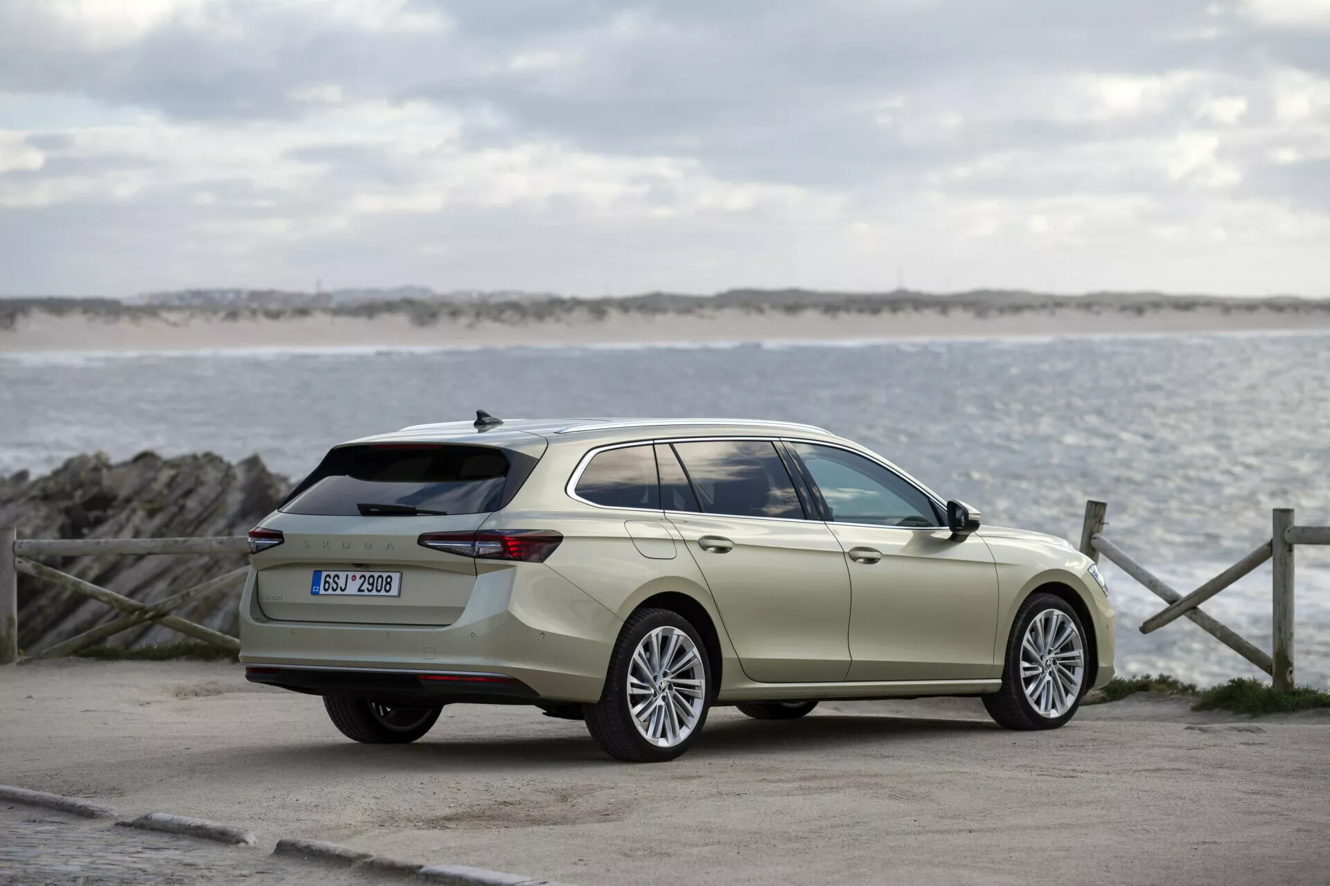2024 Skoda Superb Combi in outdoor setting with sea and sky backdrop, showcasing full side profile, large alloy wheels, body-colored door handles and mirrors, chrome window trim, extended roofline, aerodynamic body contours, and roof rails