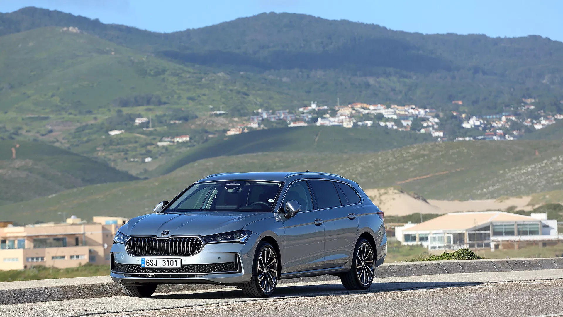 2024 Skoda Superb Combi in a three-quarter front view on a road with a hilly landscape background, featuring a prominent grille, LED headlamps, and panoramic sunroof