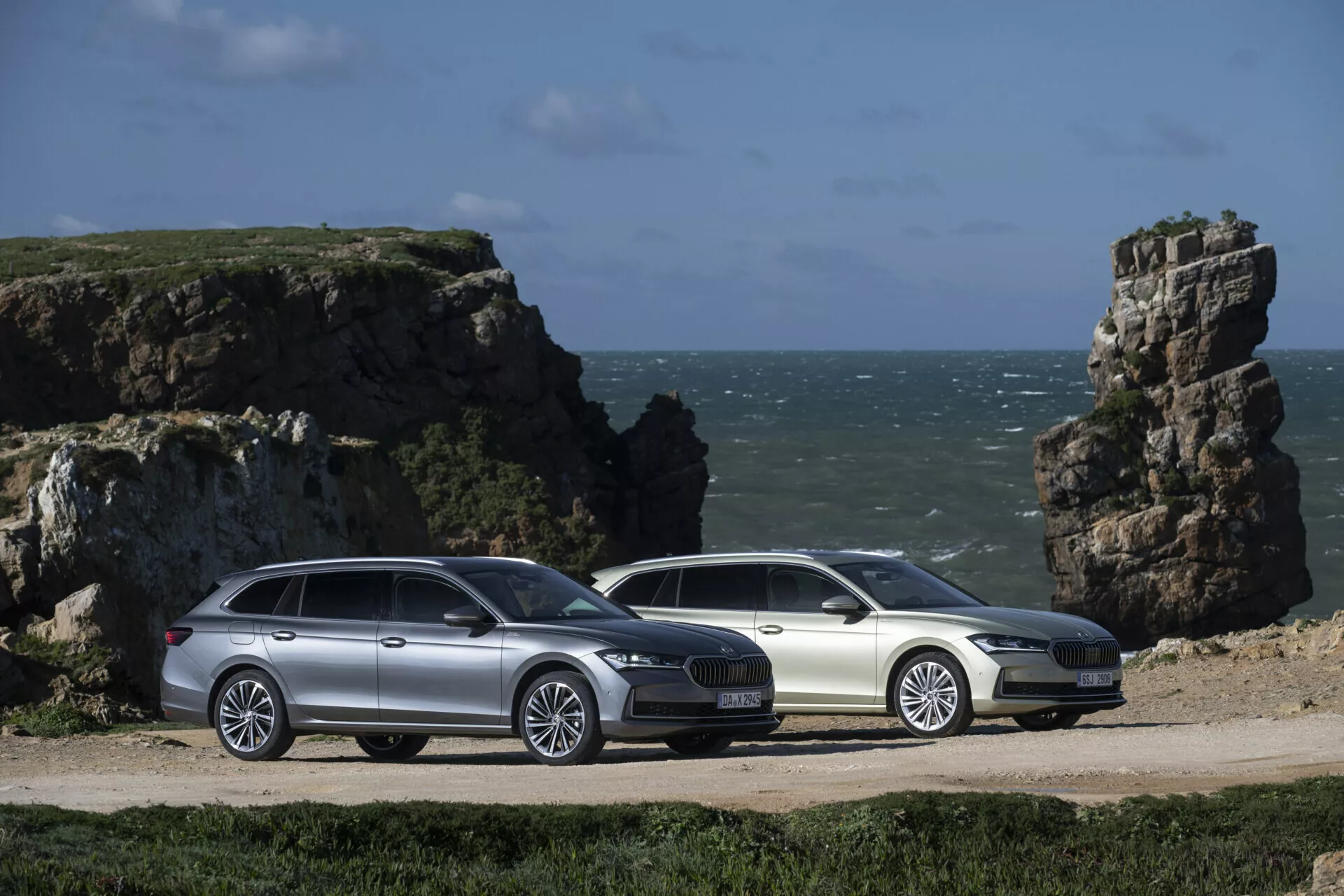 2024 Skoda Superb Combi station wagons in silver and white, displayed side by side in a three-quarter view showcasing their aerodynamic profiles, alloy wheels, and integrated roof rails, set against a coastal backdrop with rocky outcrops.