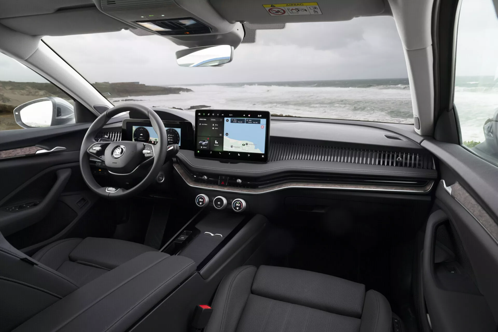 2024 Skoda Superb Combi interior showing the dashboard from the rear seat perspective, highlighting multifunction steering wheel, horizontal touchscreen infotainment system, decorative dashboard trim, dark leather seats, and panoramic roof details.