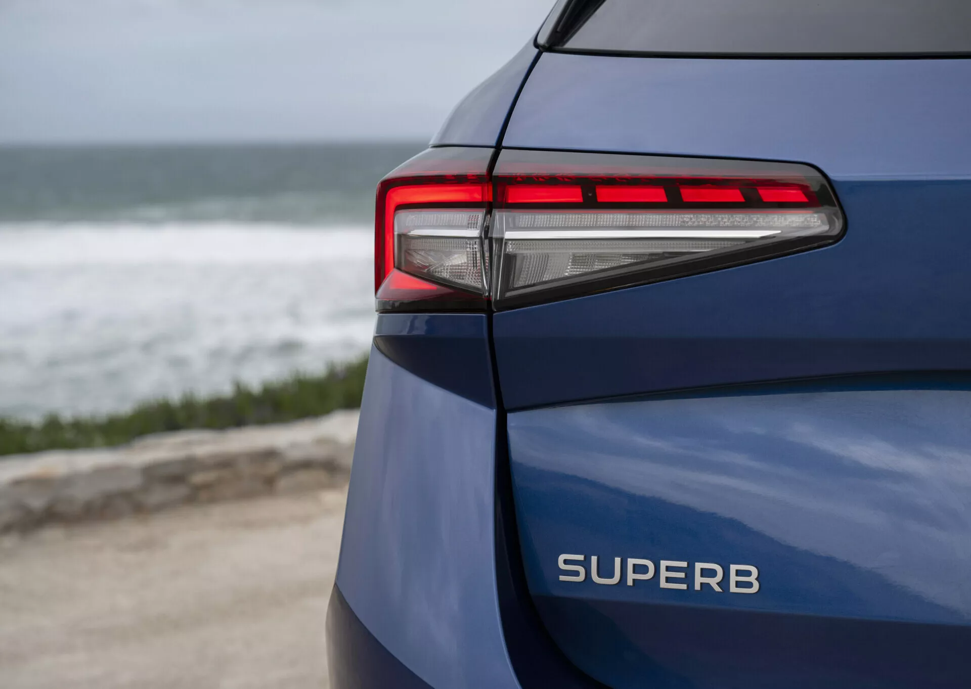 2024 Škoda Superb Combi in blue, showcasing its rear left side, distinctly displaying the SUPERB model name, LED taillight with modern horizontal lines and sharp angles against a coastal backdrop