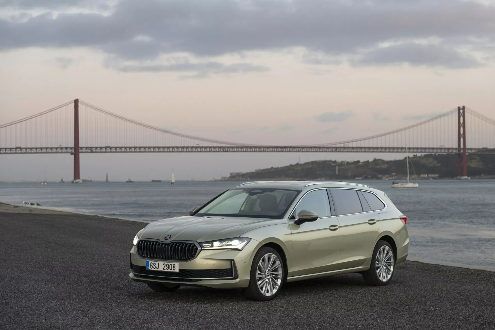 2024 Skoda Superb Combi showcased in a three-quarter front view highlighting the sleek hatchback design, chrome-finished front grille, LED headlights, and alloy wheels set against a scenic backdrop with a water body and bridge at dusk or dawn