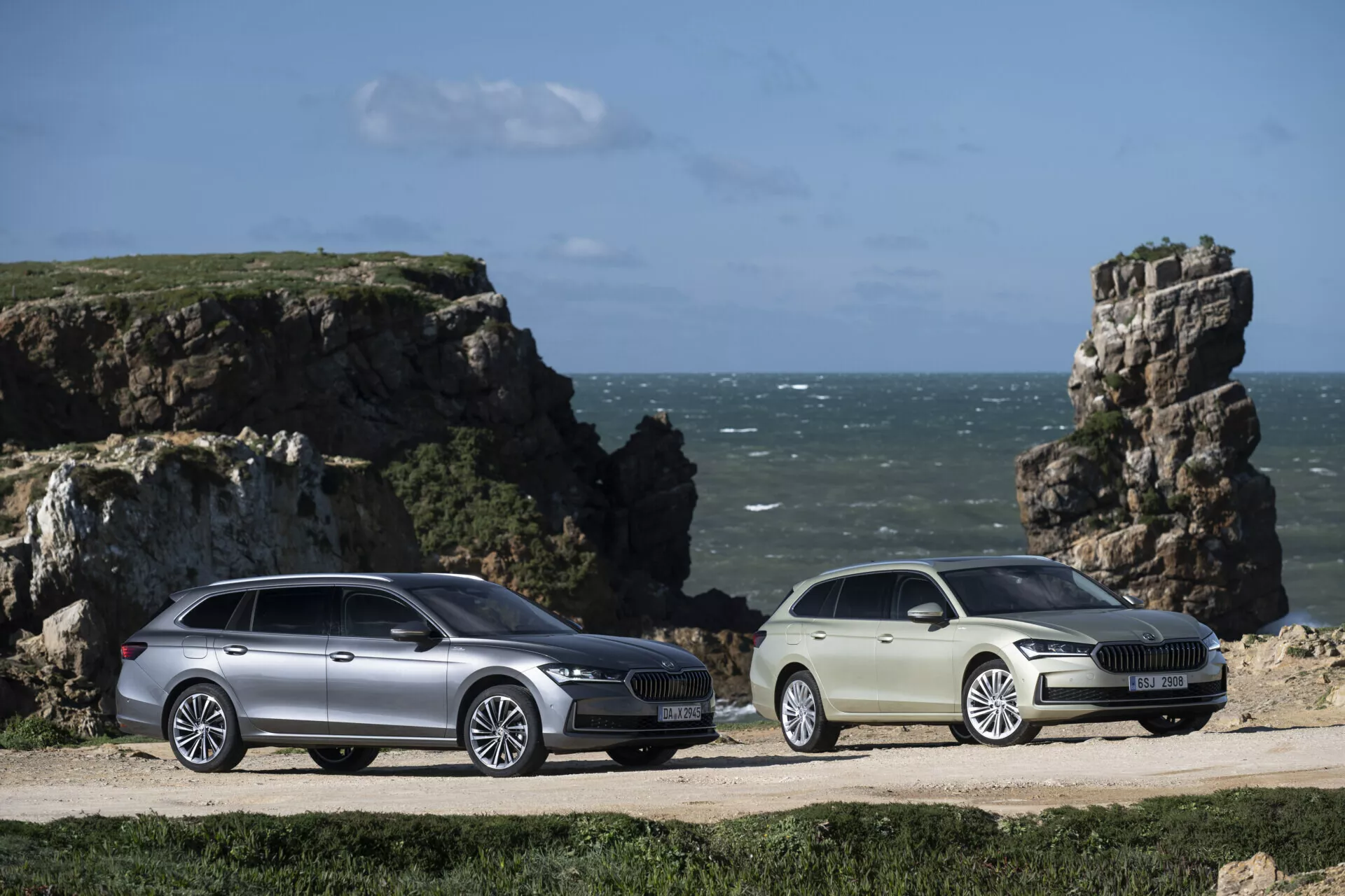 2024 Skoda Superb Combi and sedan models parked side by side on a coastal dirt path with a rocky seaside cliff and a blue sky background, featuring extended cargo space, aerodynamic design, LED headlights, and alloy wheels, emphasizing their adventurous and lifestyle-oriented appeal.
