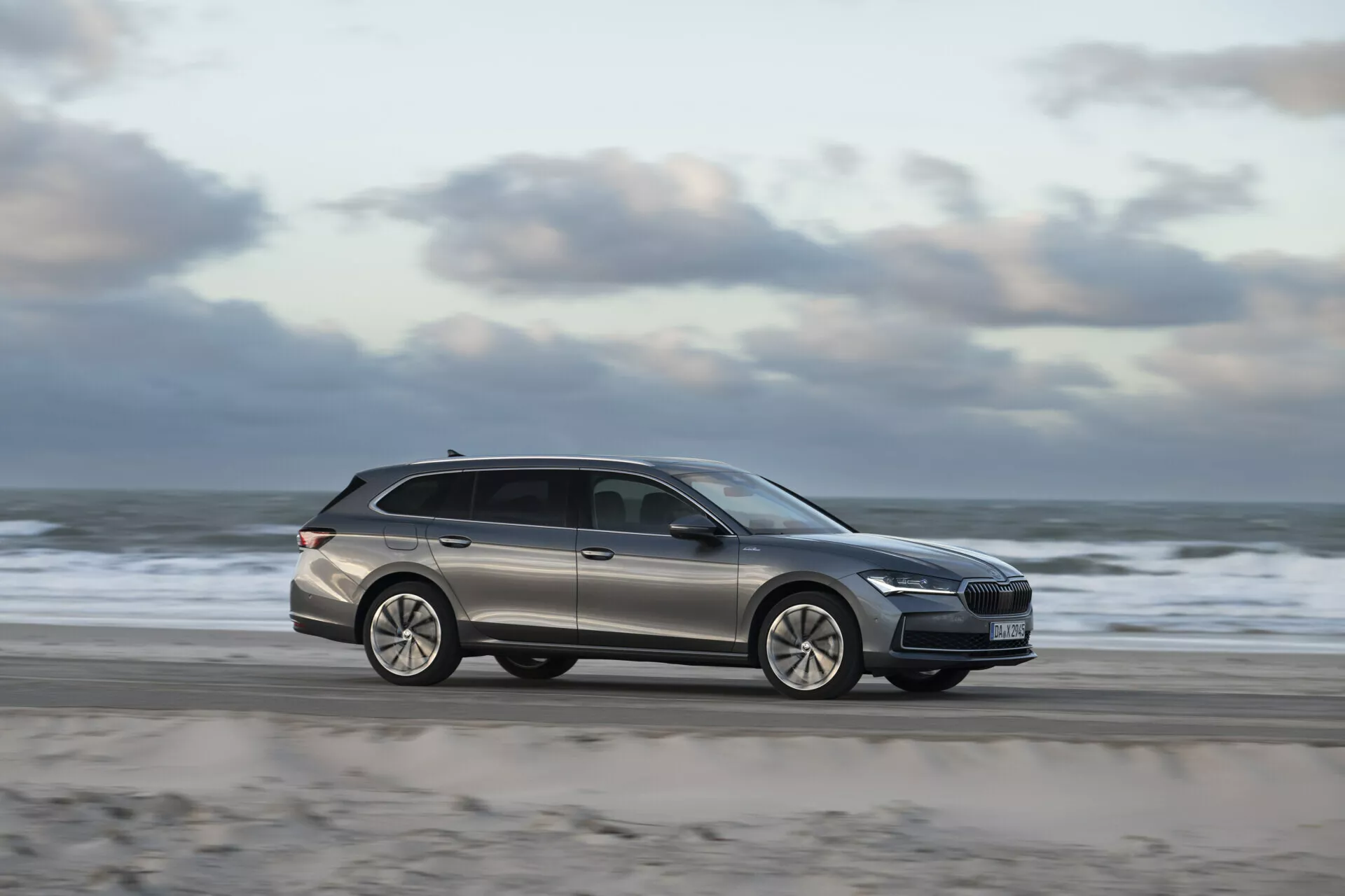 2024 Skoda Superb Combi station wagon driving on a beachside road, showcasing modern design with side profile view, motion blur indicating speed, alloy wheels, and roof rails for functionality.